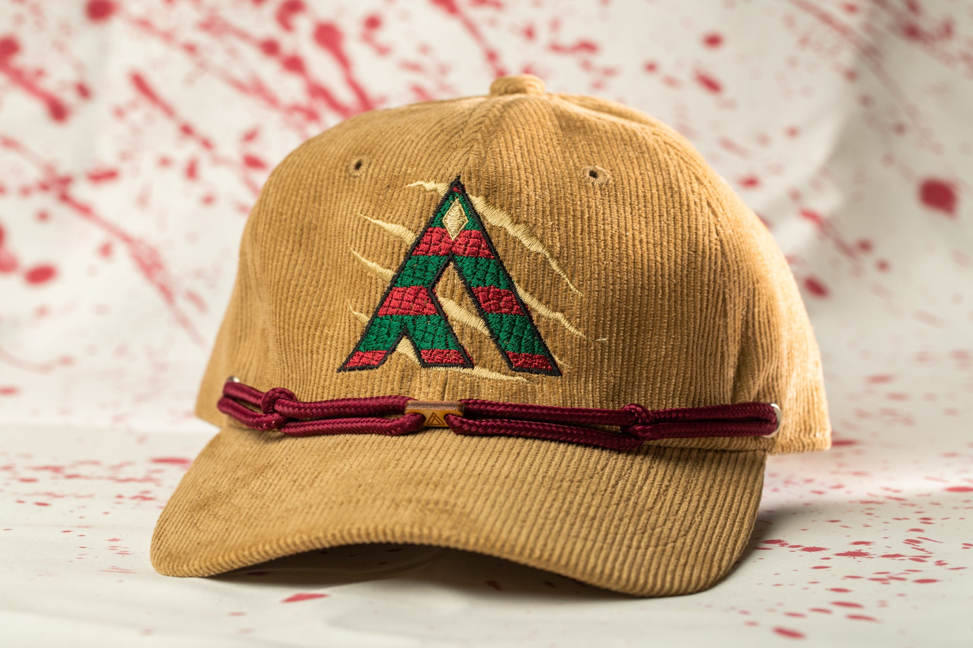 A beige baseball cap with a red and green embroidered logo on the front, sitting on a white surface with red splatters in the background.