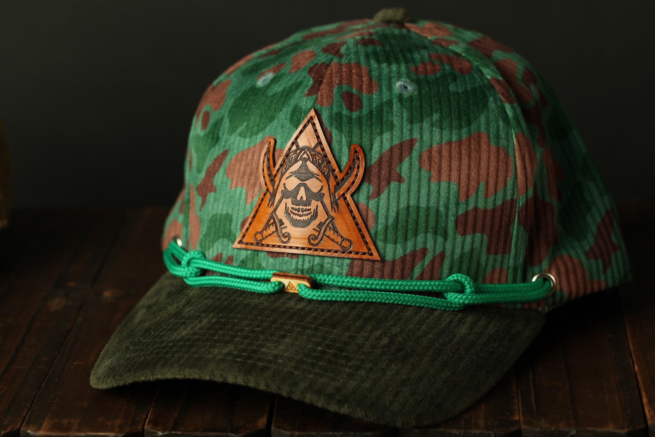 A camouflage baseball cap with a skull and crossbones design on the front, sitting on a wooden surface.