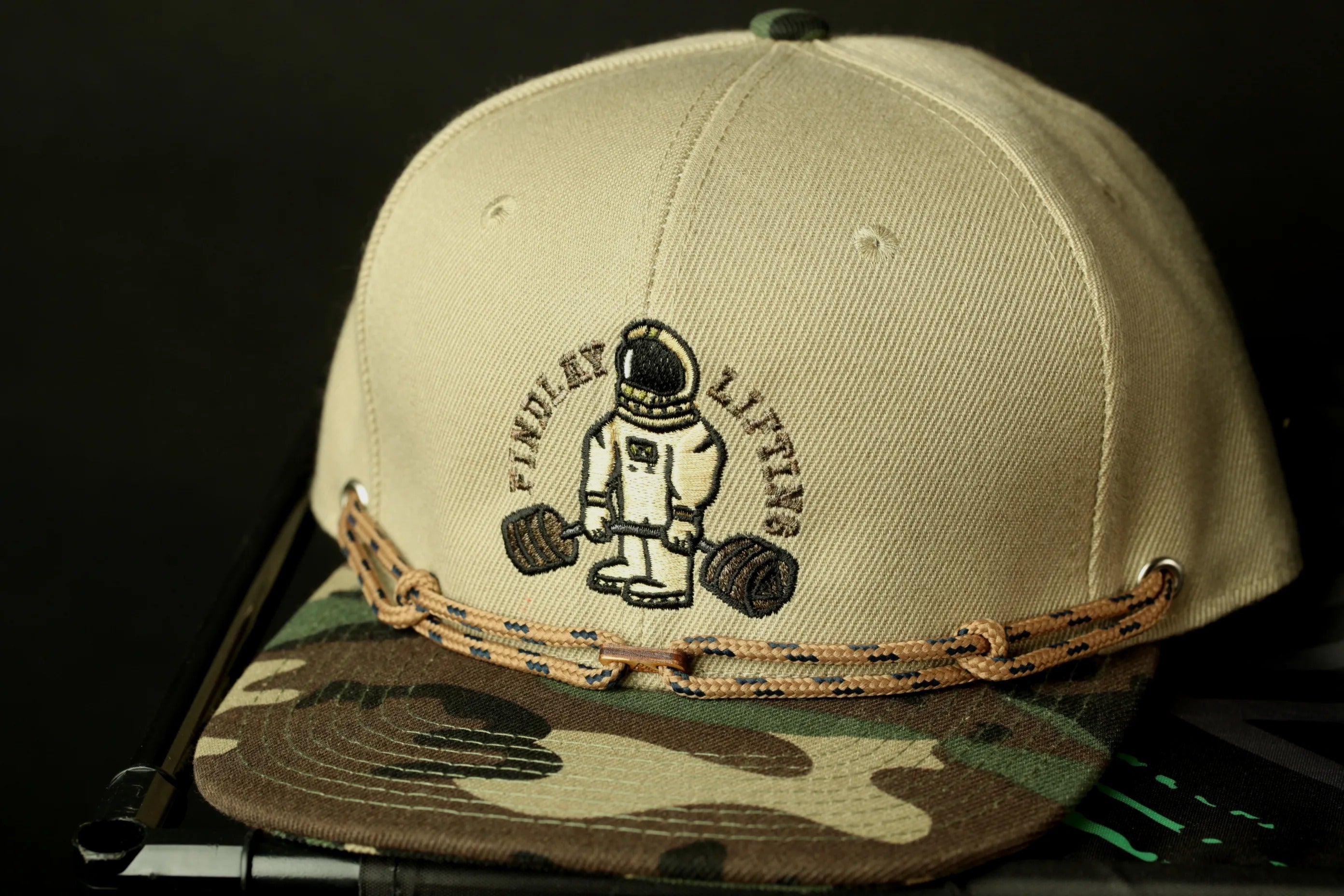 A beige baseball cap with a camouflage pattern and a graphic of a person lifting weights.