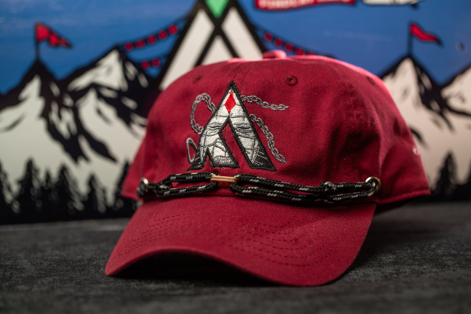 A red baseball cap with a black and white logo on the front, placed on a gray surface against a background of mountains and flags.