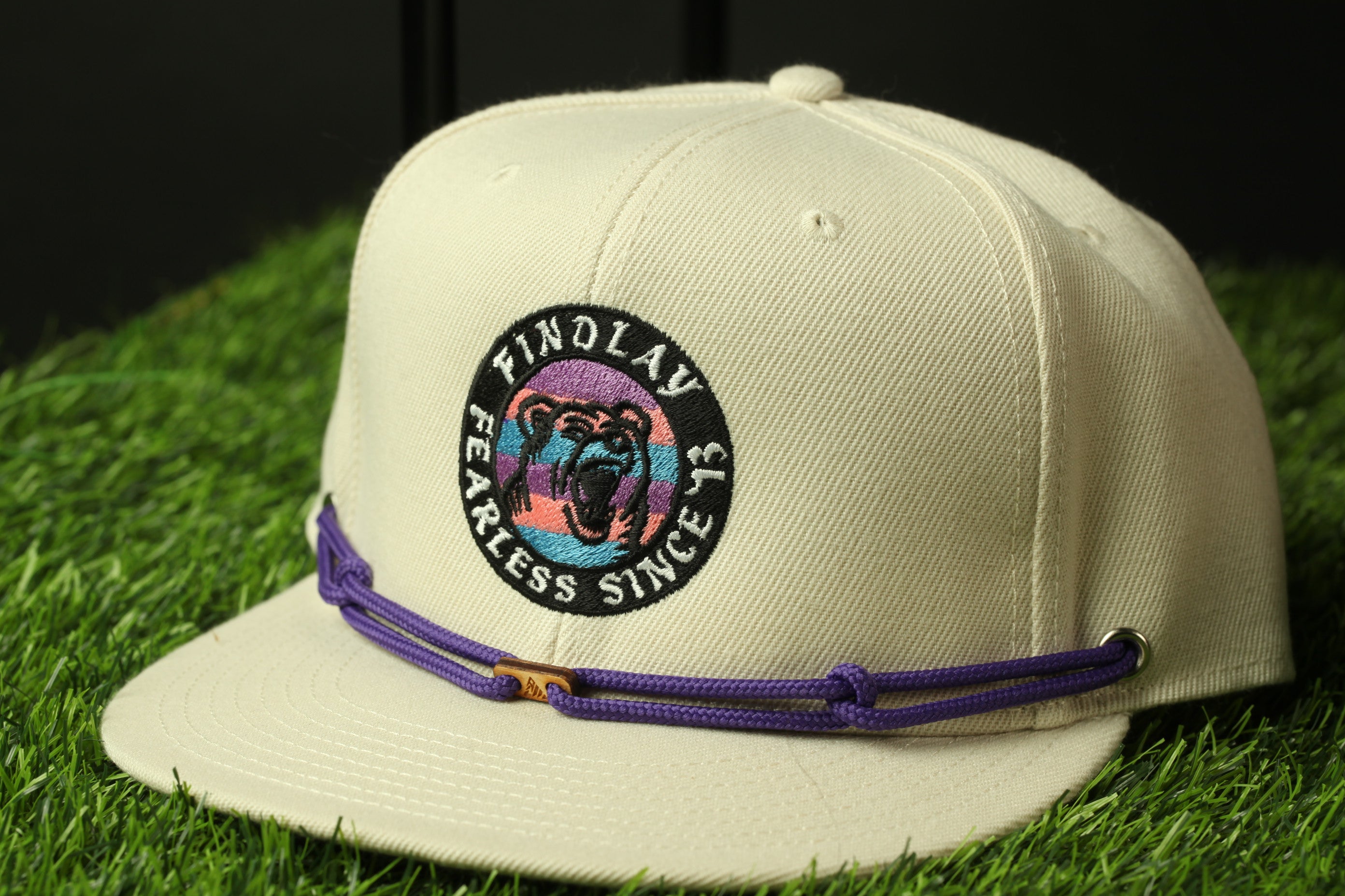 A beige baseball cap with a purple band and a circular logo on the front, placed on a grassy surface.
