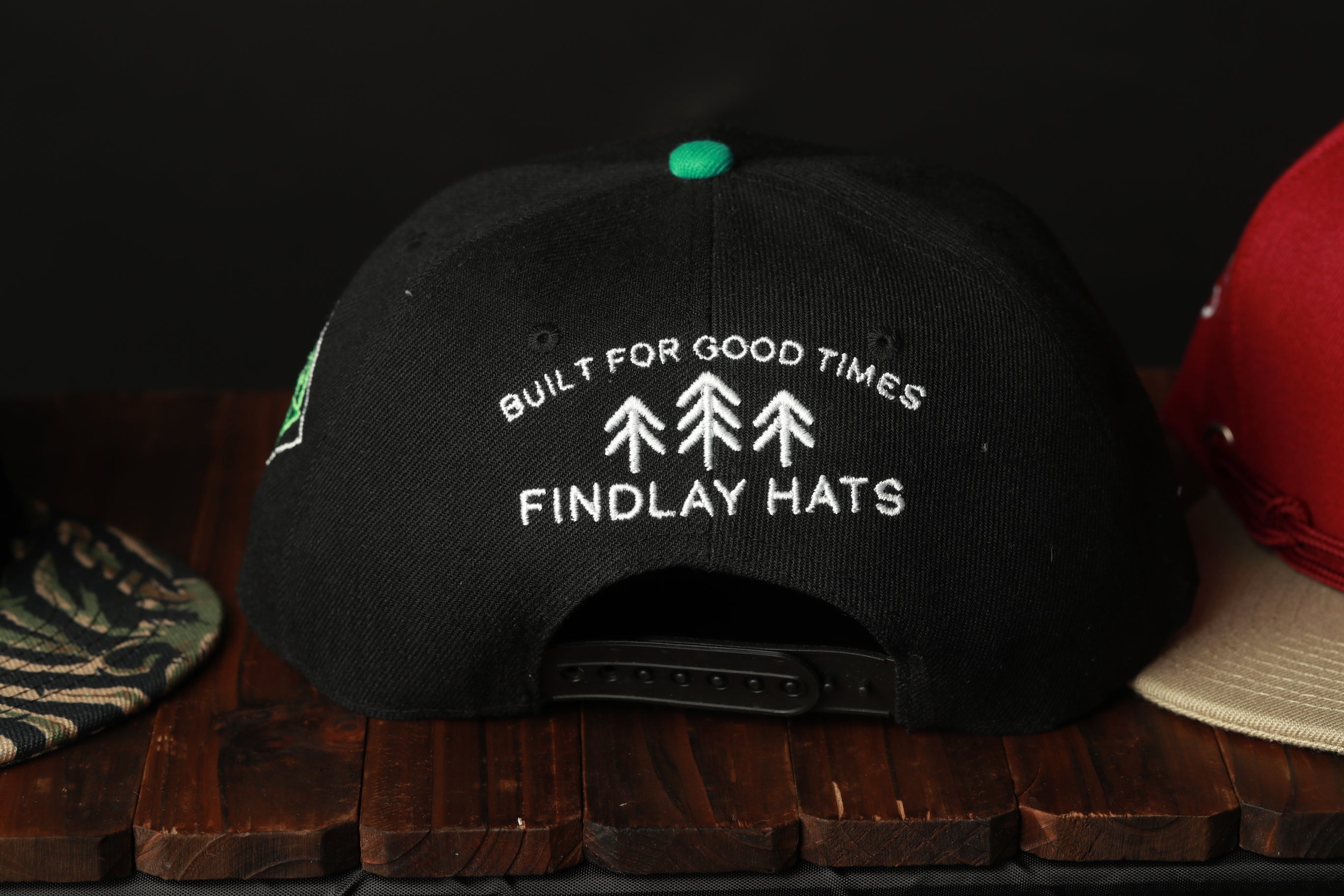 A black baseball cap with white text and a green tip, sitting on a wooden surface.