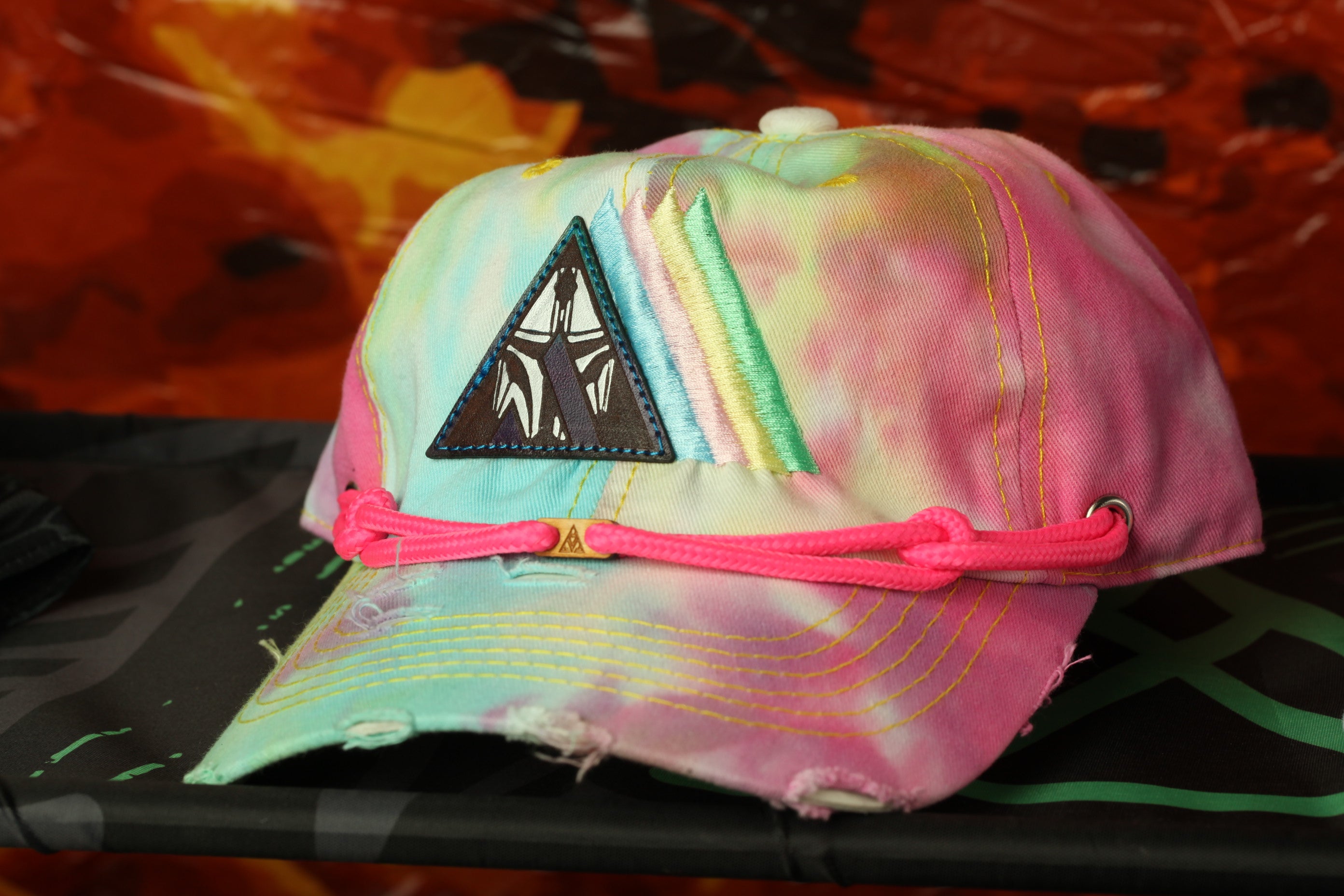 A colorful, tie-dye baseball cap with a black patch and pink braided strap is displayed against a red background.