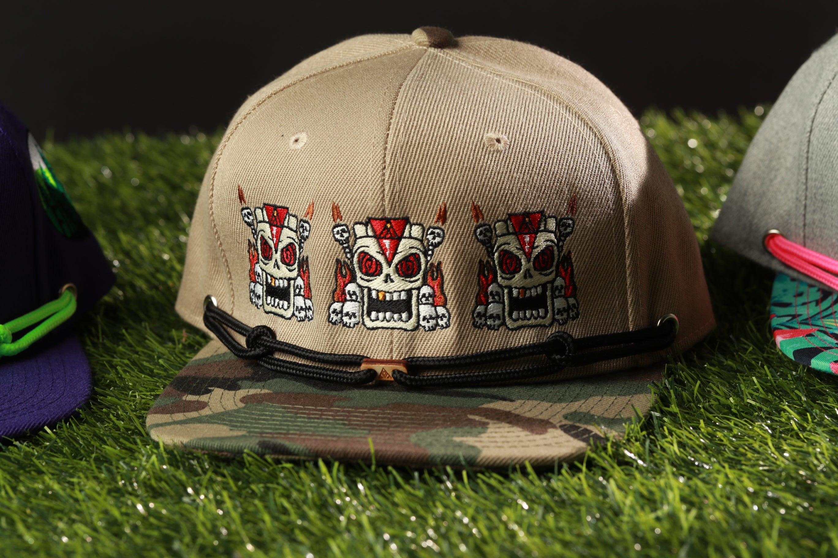 A beige baseball cap with a camouflage pattern and three skull designs on it, placed on a grassy surface.