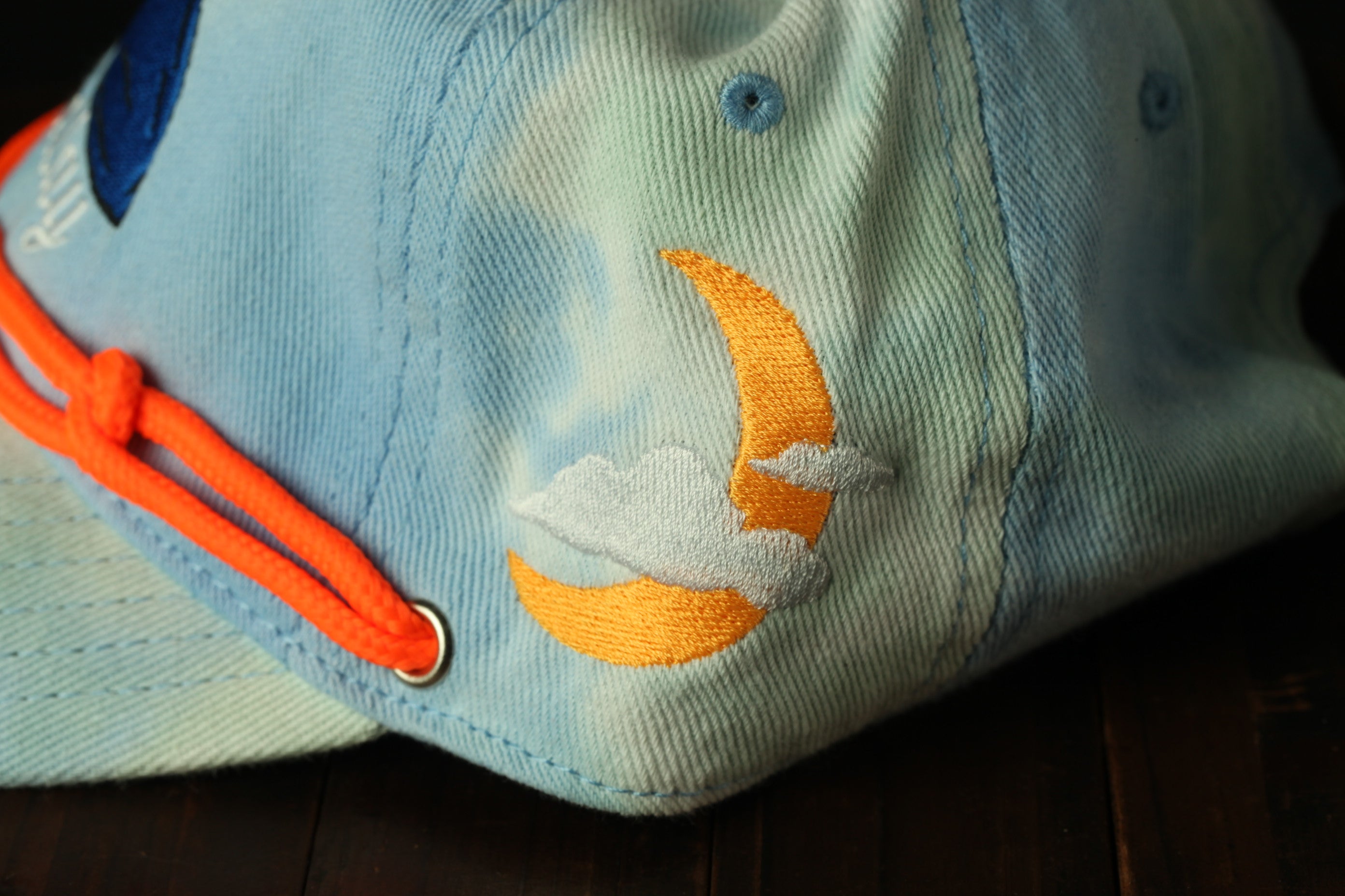 A close-up view of a blue baseball cap with a crescent moon and clouds embroidered on it, along with an orange string or strap.