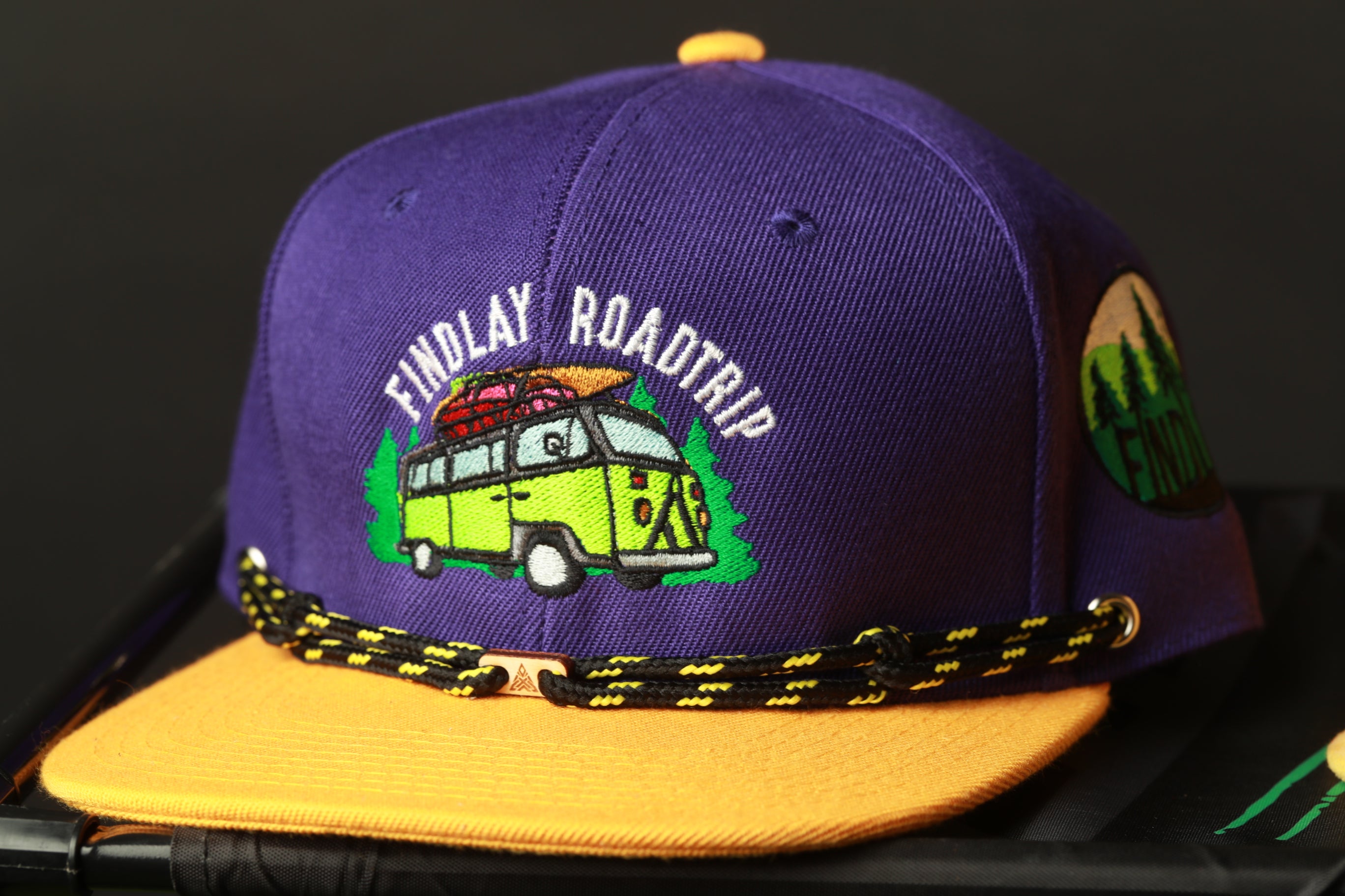 A purple baseball cap with a yellow brim and a graphic of a van on it, along with the text "Findlay Roadtrip" printed on it.