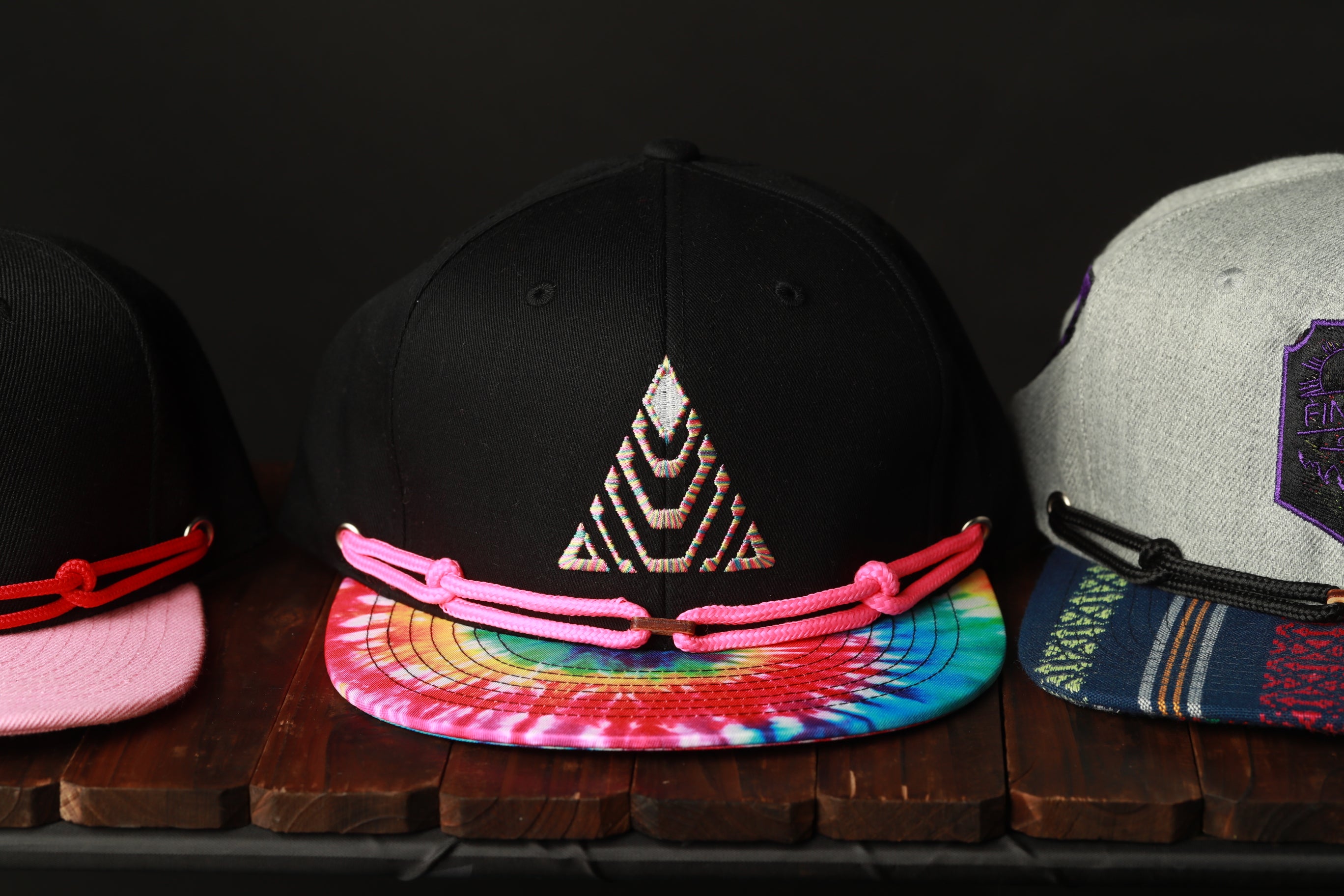 Three colorful baseball caps are displayed on a wooden surface, with one black cap featuring a tie-dye pattern and two gray caps with different patterns.