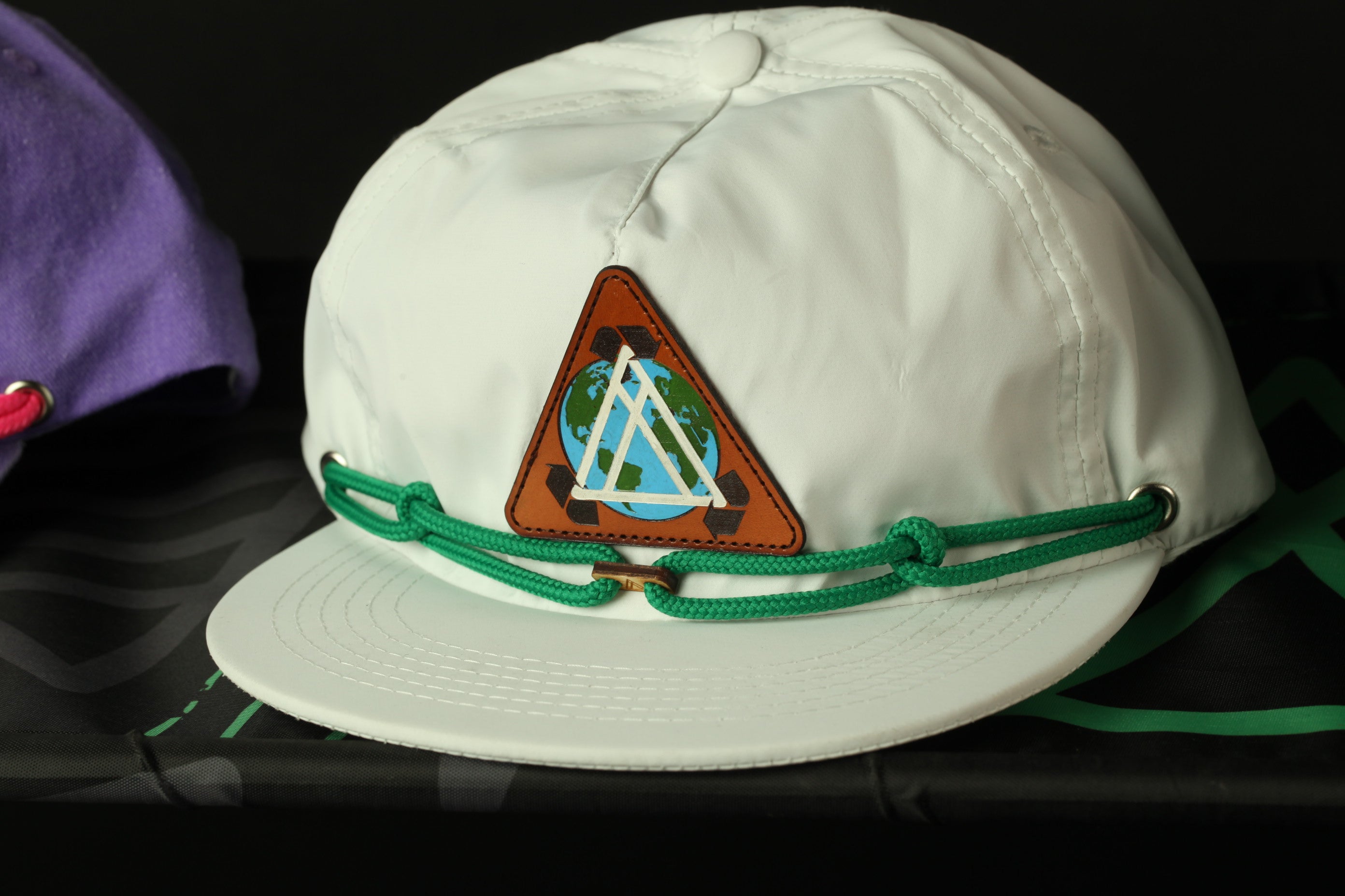 A white baseball cap with a triangular patch featuring a globe and a triangle design, and green strings around the brim.