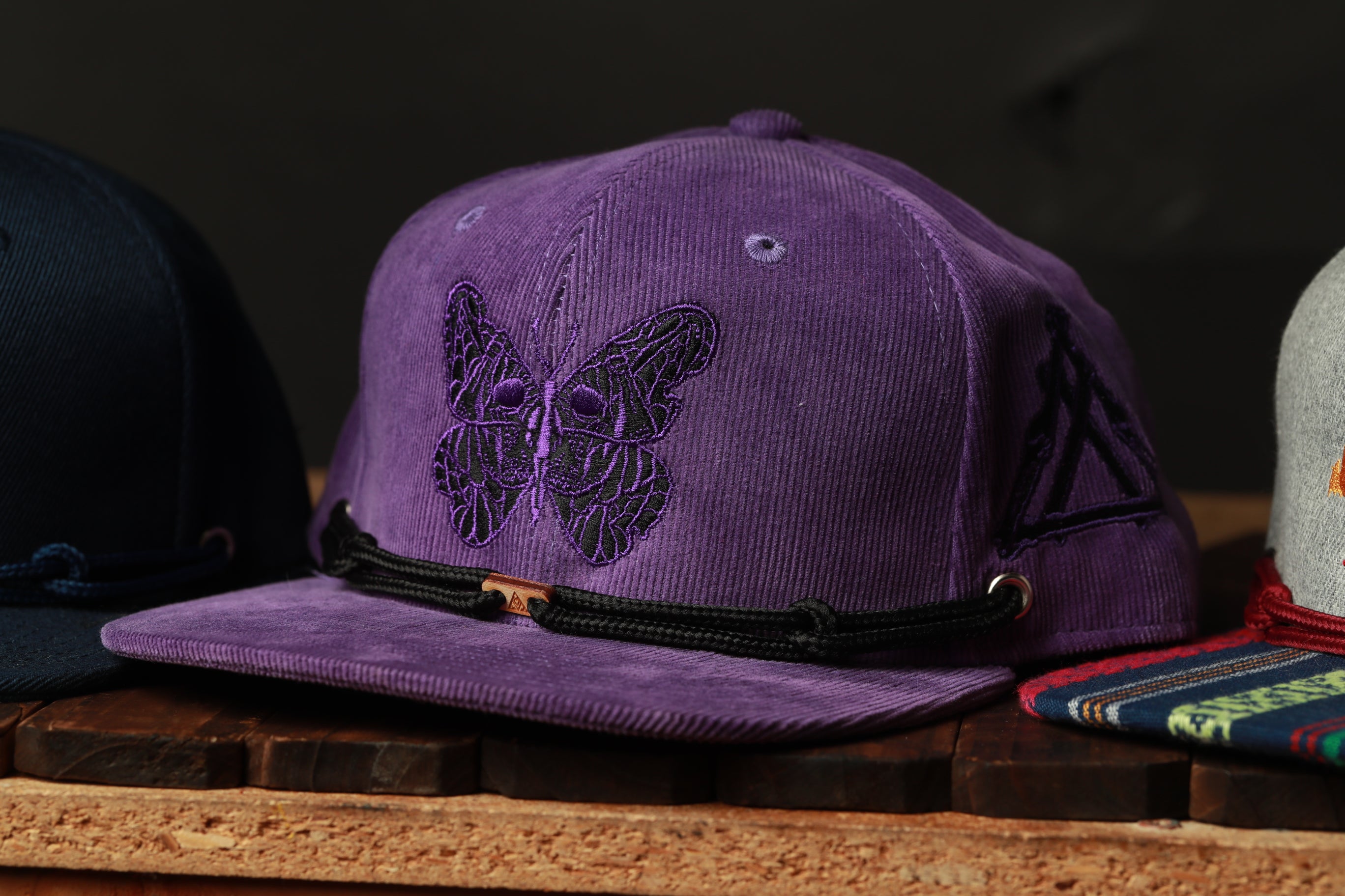 A purple baseball cap with a butterfly design is displayed on a wooden surface, with other hats visible in the background.