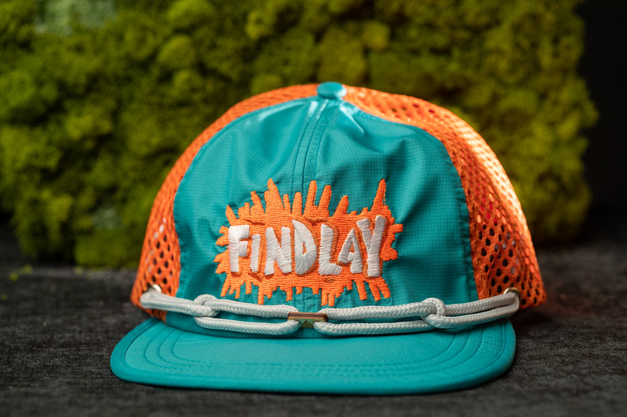 A turquoise baseball cap with orange splatter designs and the word "FINDlay" printed on it, placed on a dark surface against a green mossy background.