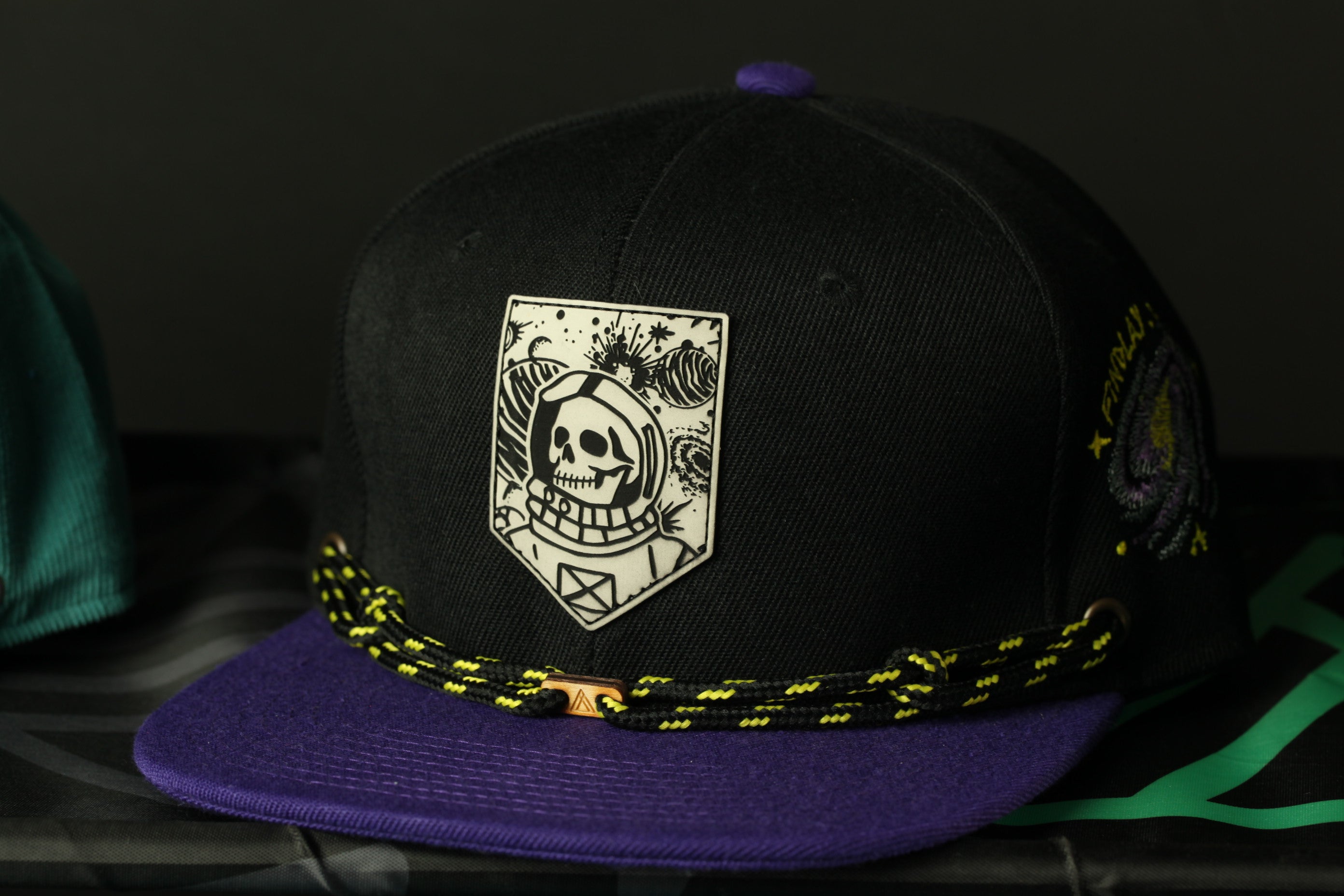 A black baseball cap with a purple brim and a skull and crossbones design on the front, along with a yellow and black braided strap.