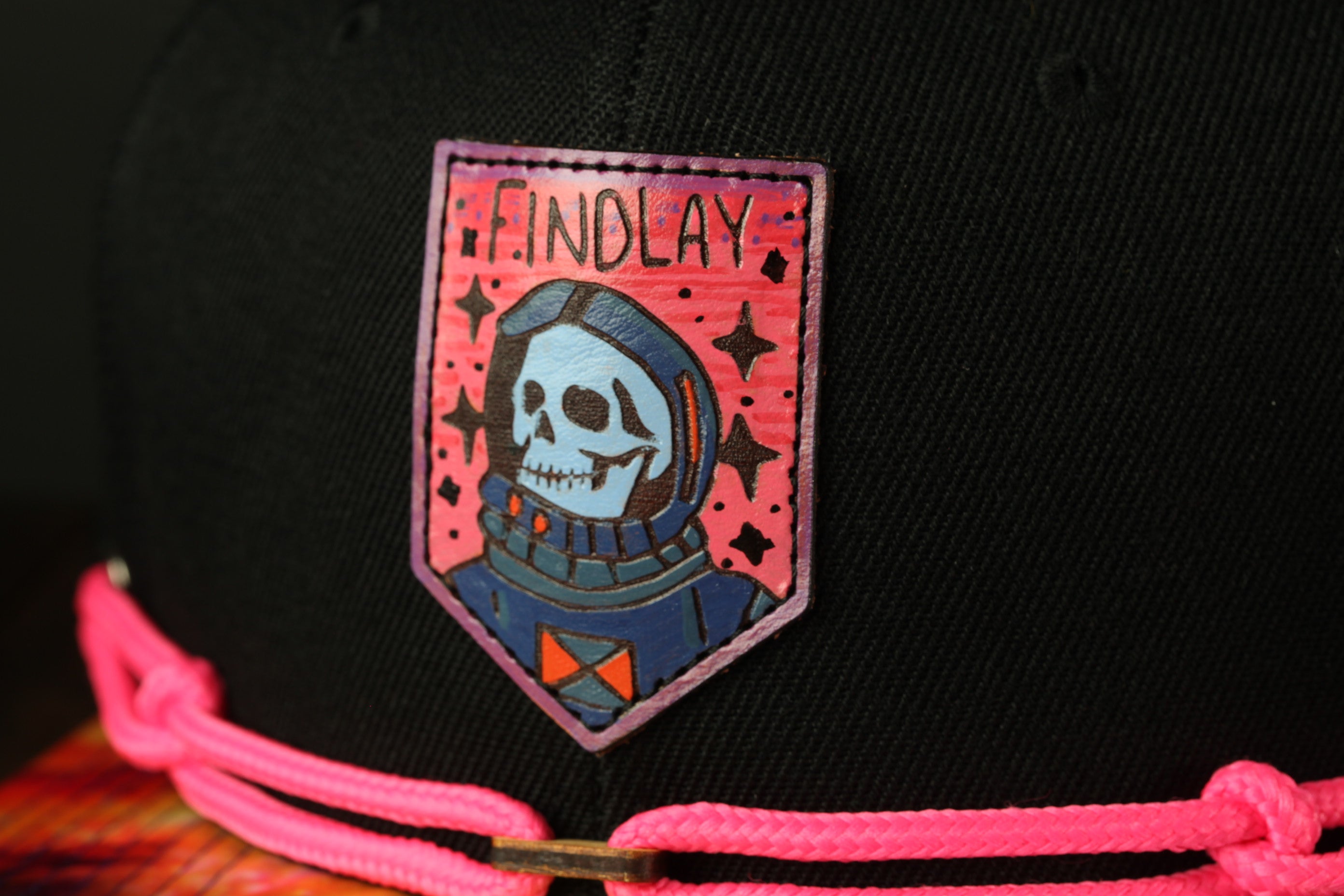 A close-up of a black baseball cap with a colorful patch featuring a skull wearing a spacesuit and the word "FINDlay" written on it.