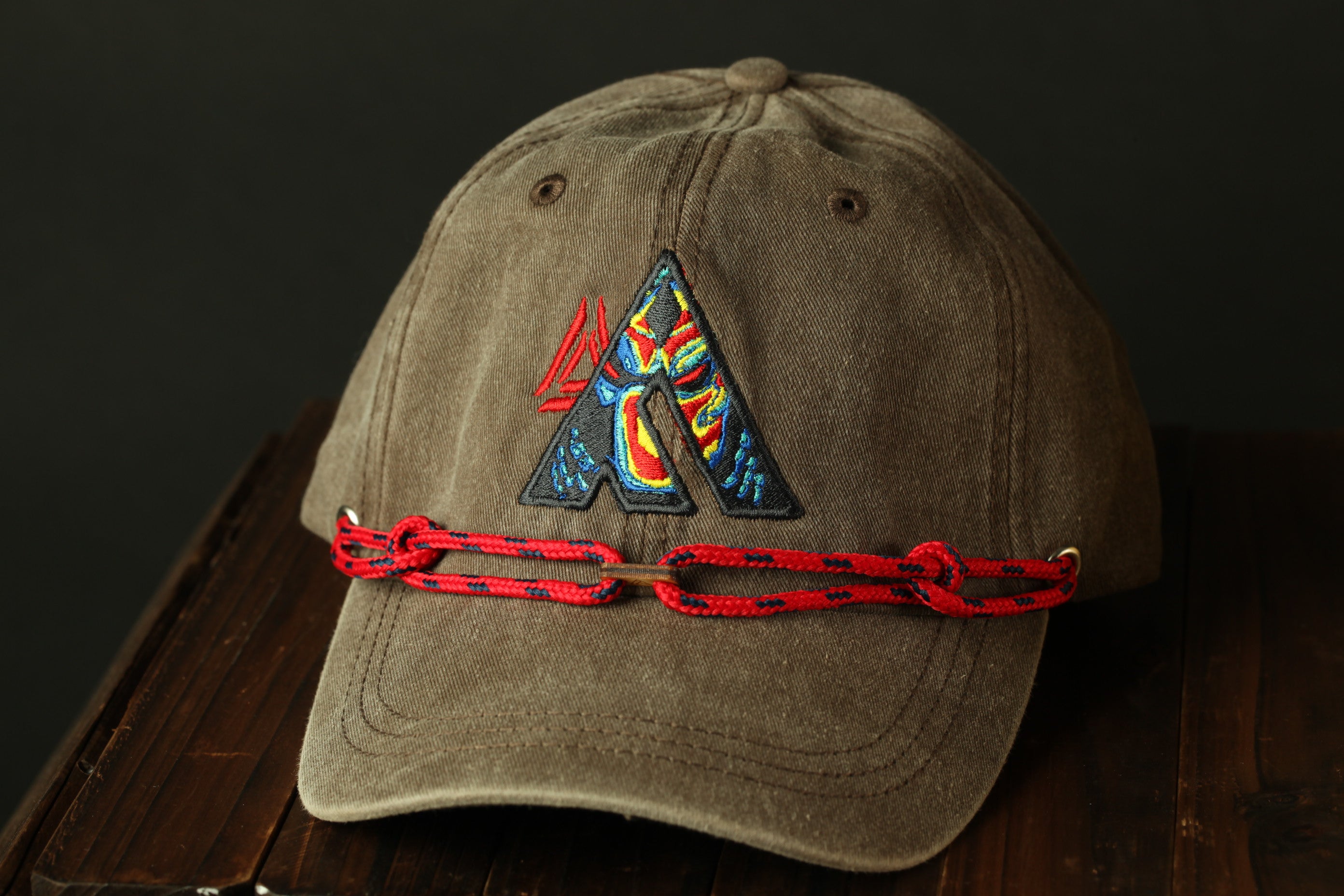 A brown baseball cap with a colorful, embroidered logo on the front, and a red string or chain attached to it.