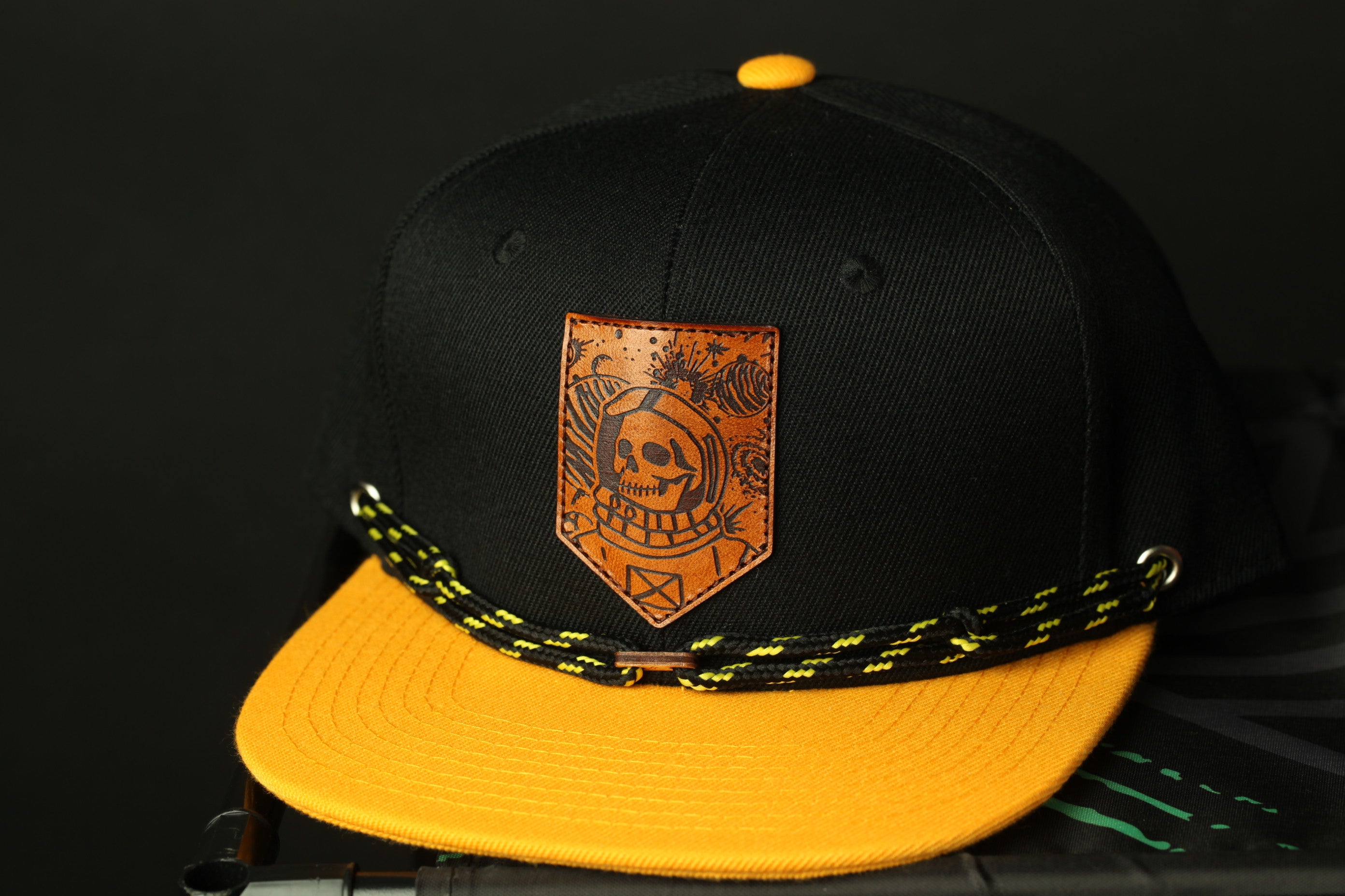 A black baseball cap with a yellow brim and a distinctive orange patch featuring a skull design.