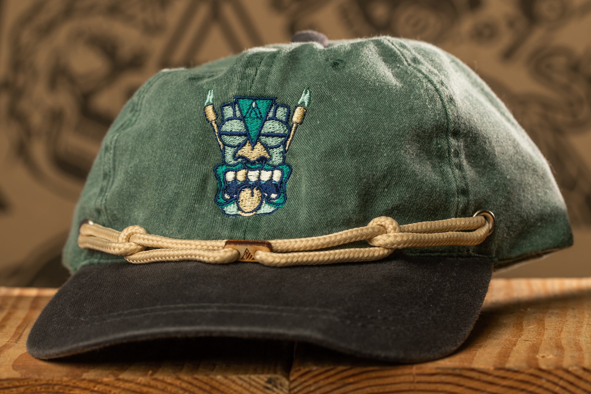 A green baseball cap with a tiki mask embroidered on the front, featuring a rope strap.