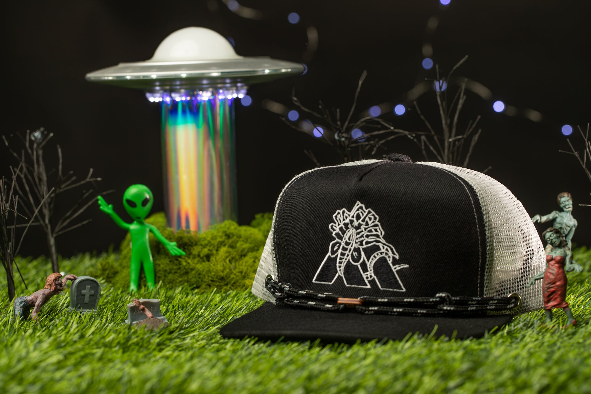 a green alien figurine, a black and white baseball cap with a logo, a miniature alien spacecraft, and a miniature landscape with a grassy area, trees, and a skull.