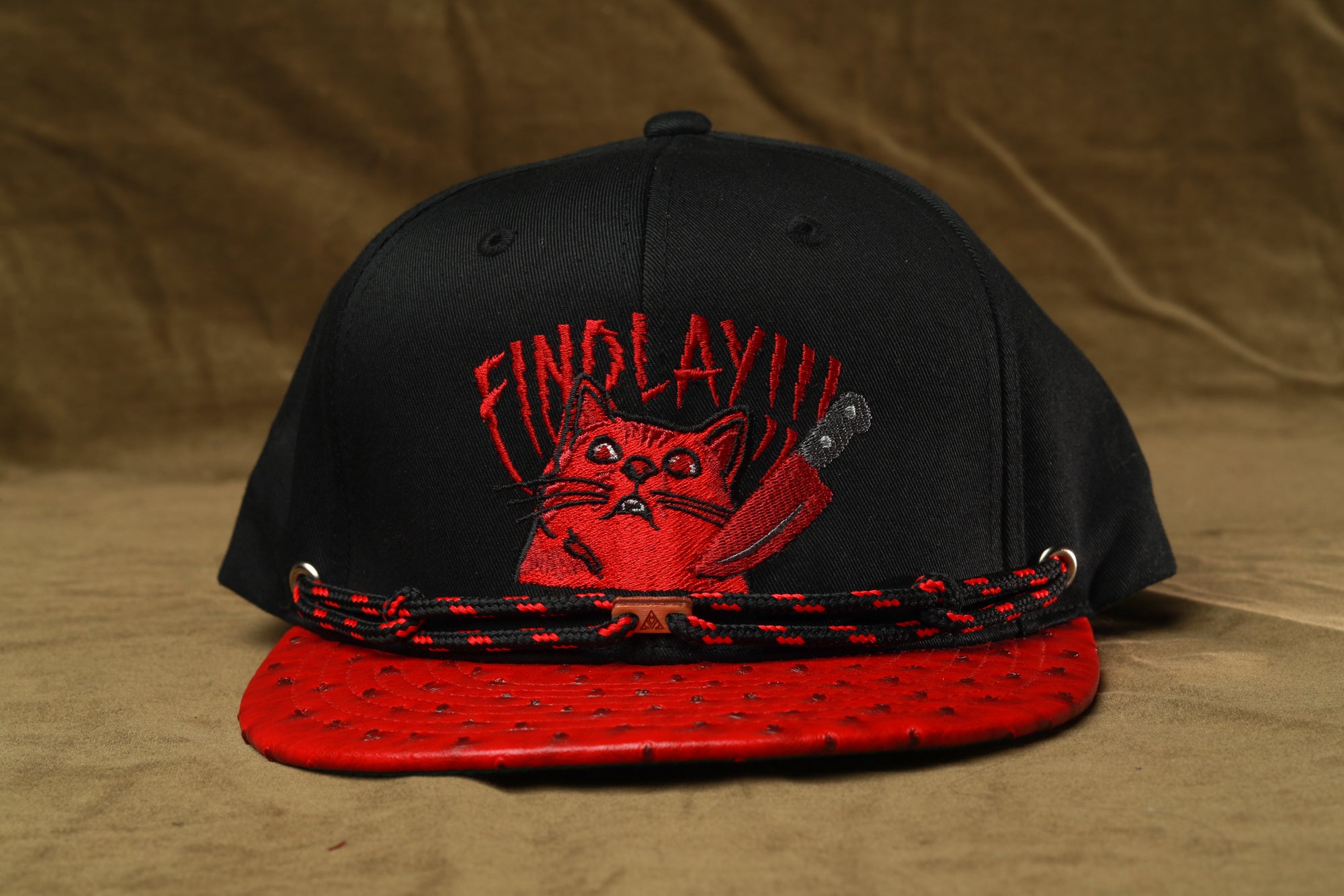 A black baseball cap with a red cat embroidered on it, and a red and black braided strap.