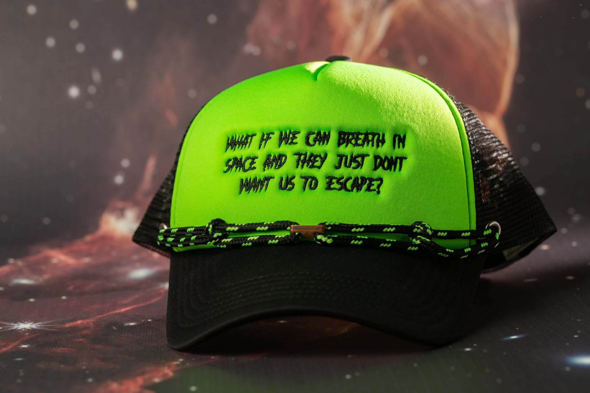 A green baseball cap with black text on it, placed on a surface with a space-themed background.