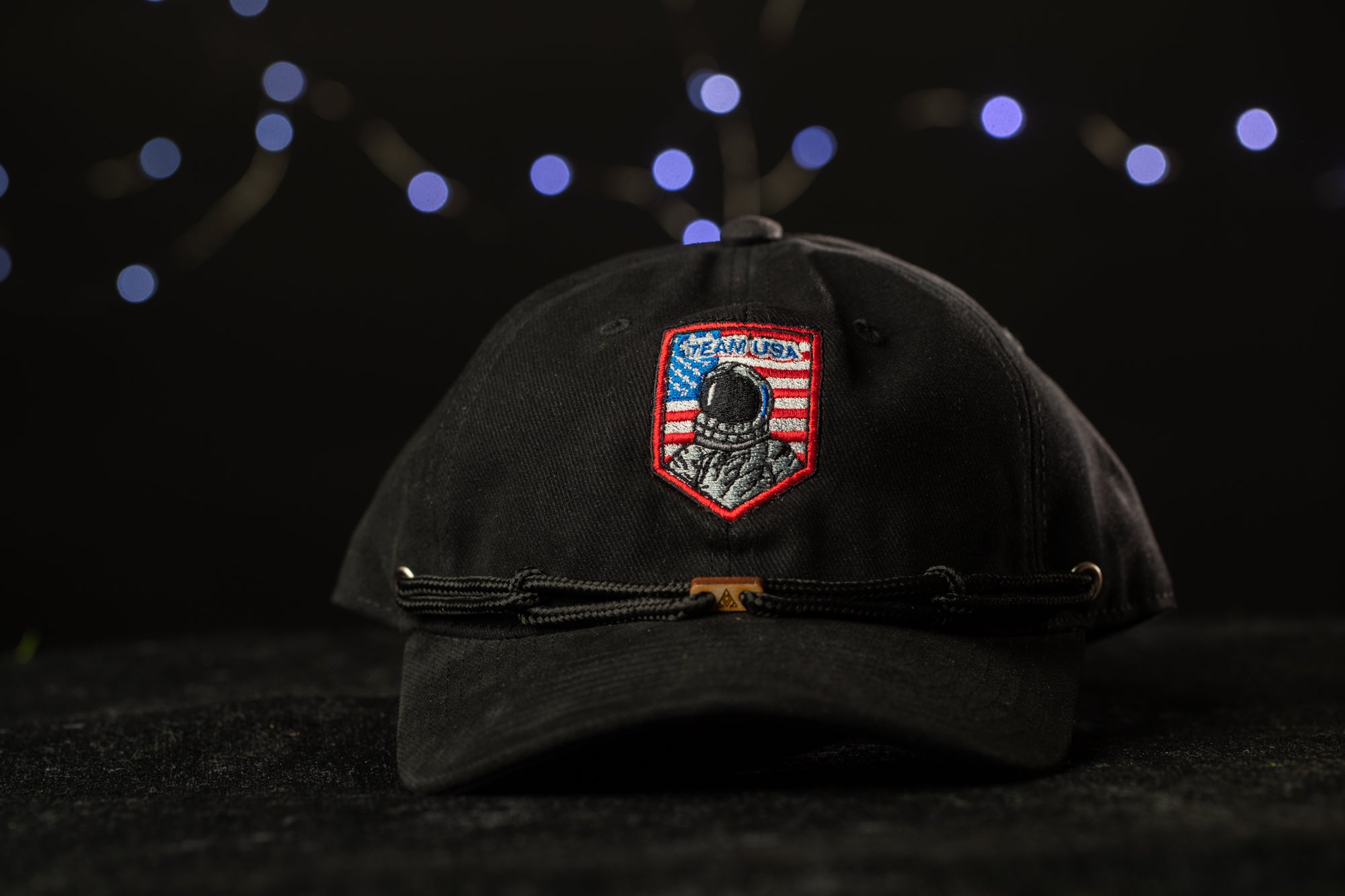 A black baseball cap with a patch featuring an astronaut and the word "USA" on it, placed on a dark surface against a background of blue lights.