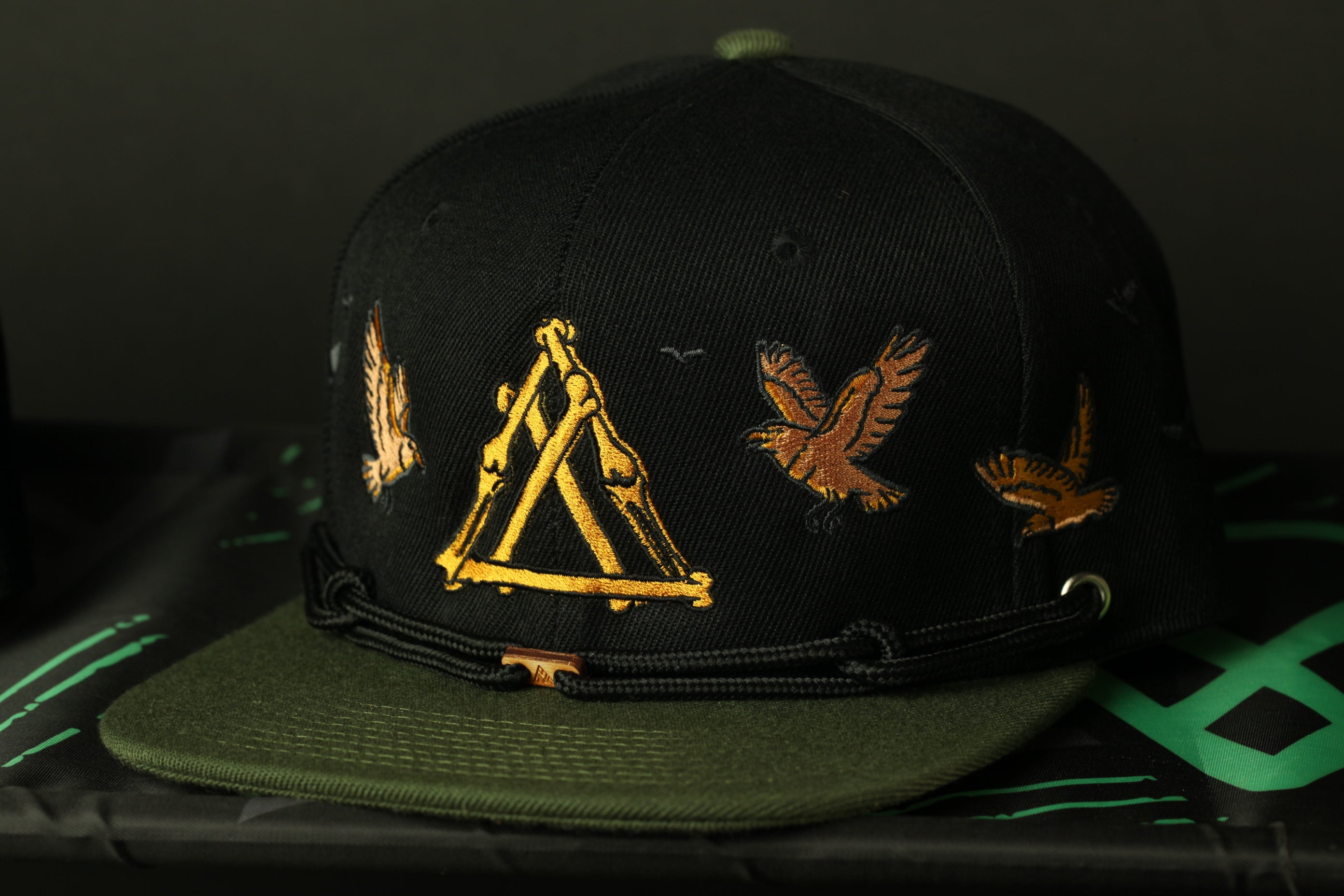 A black baseball cap with gold embroidered designs, including a triangle, a bird, and another bird, against a dark background.