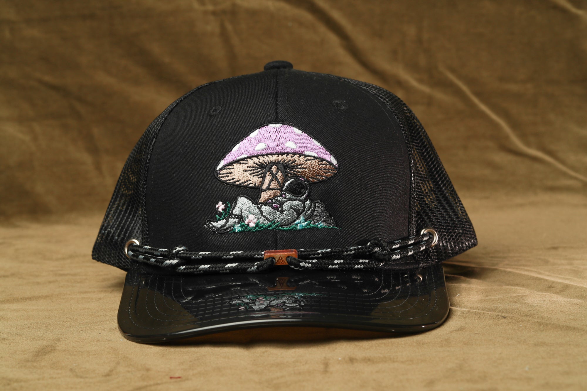 A black baseball cap with a mushroom design on the front, featuring a small figure of a person sitting under the mushroom.
