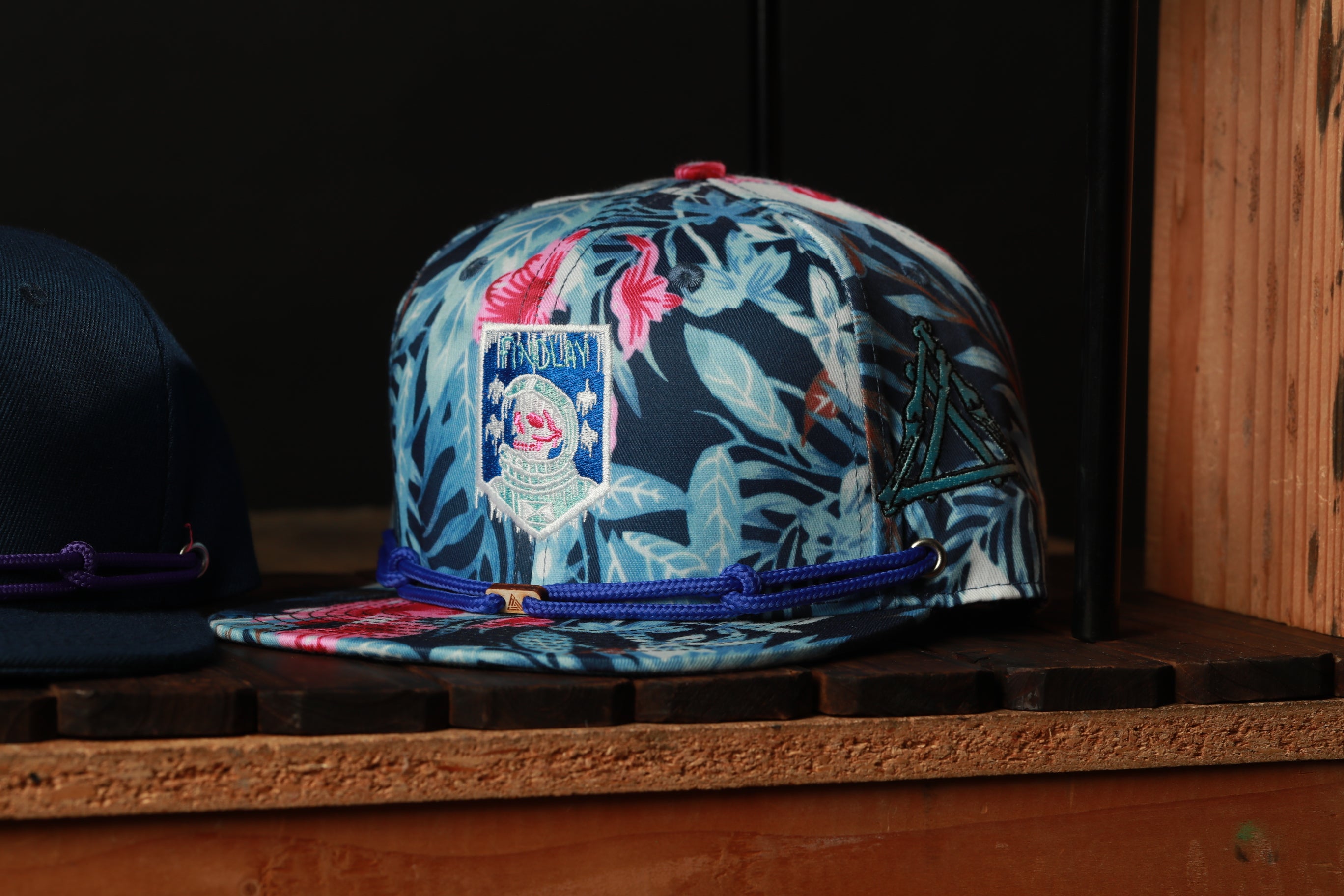 A colorful floral-patterned baseball cap with a blue brim and a small emblem on the front, displayed on a wooden shelf.