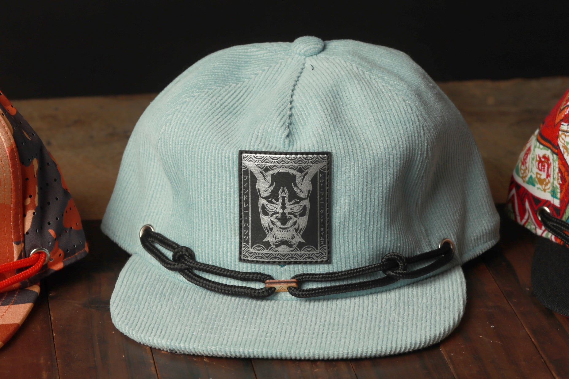 A light blue baseball cap with a black chain strap and a graphic patch on the front, sitting on a wooden surface.