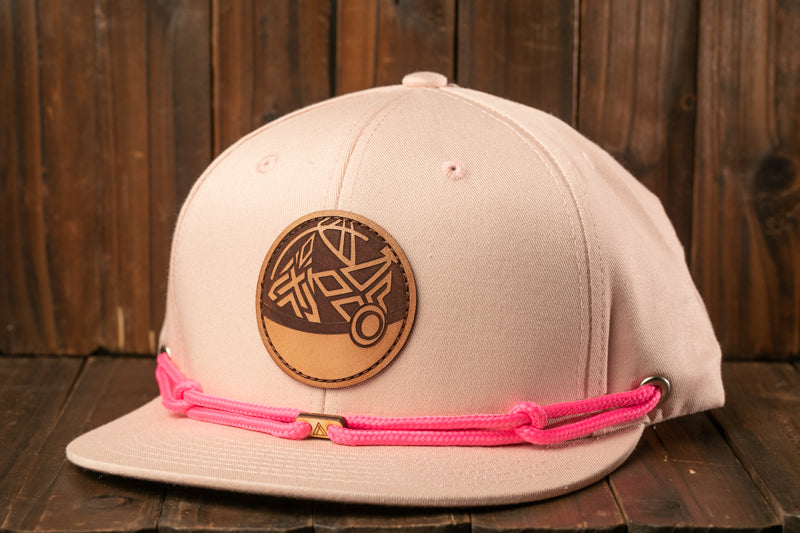 A pink baseball cap with a leather patch and pink braided strap is displayed against a wooden background.