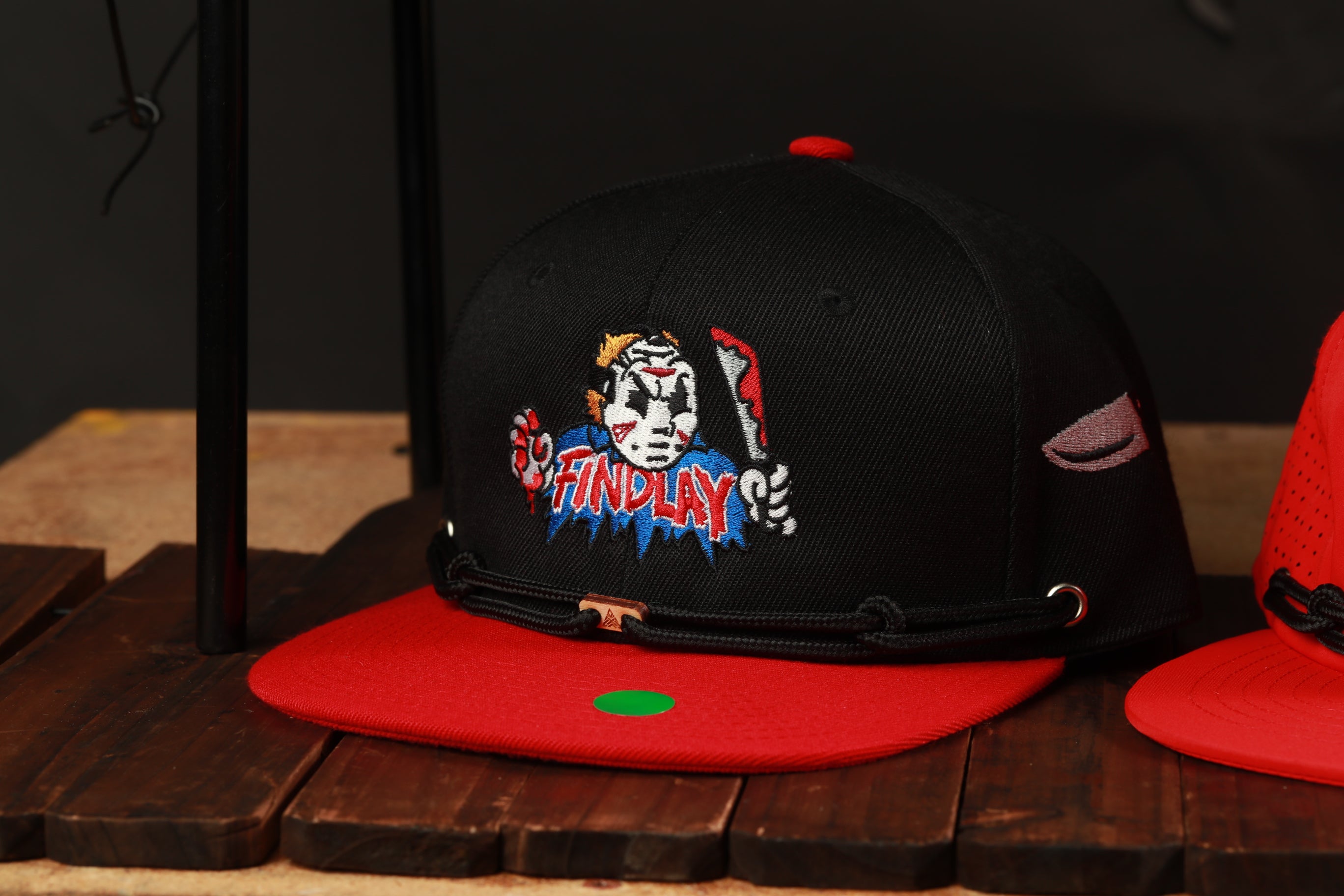 A black baseball cap with a red brim and a graphic design on the front, placed on a wooden surface.