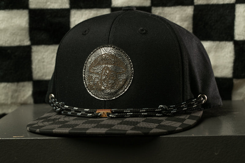 A black baseball cap with a checkered pattern and a metal emblem on the front, displayed against a black and white checkered background.