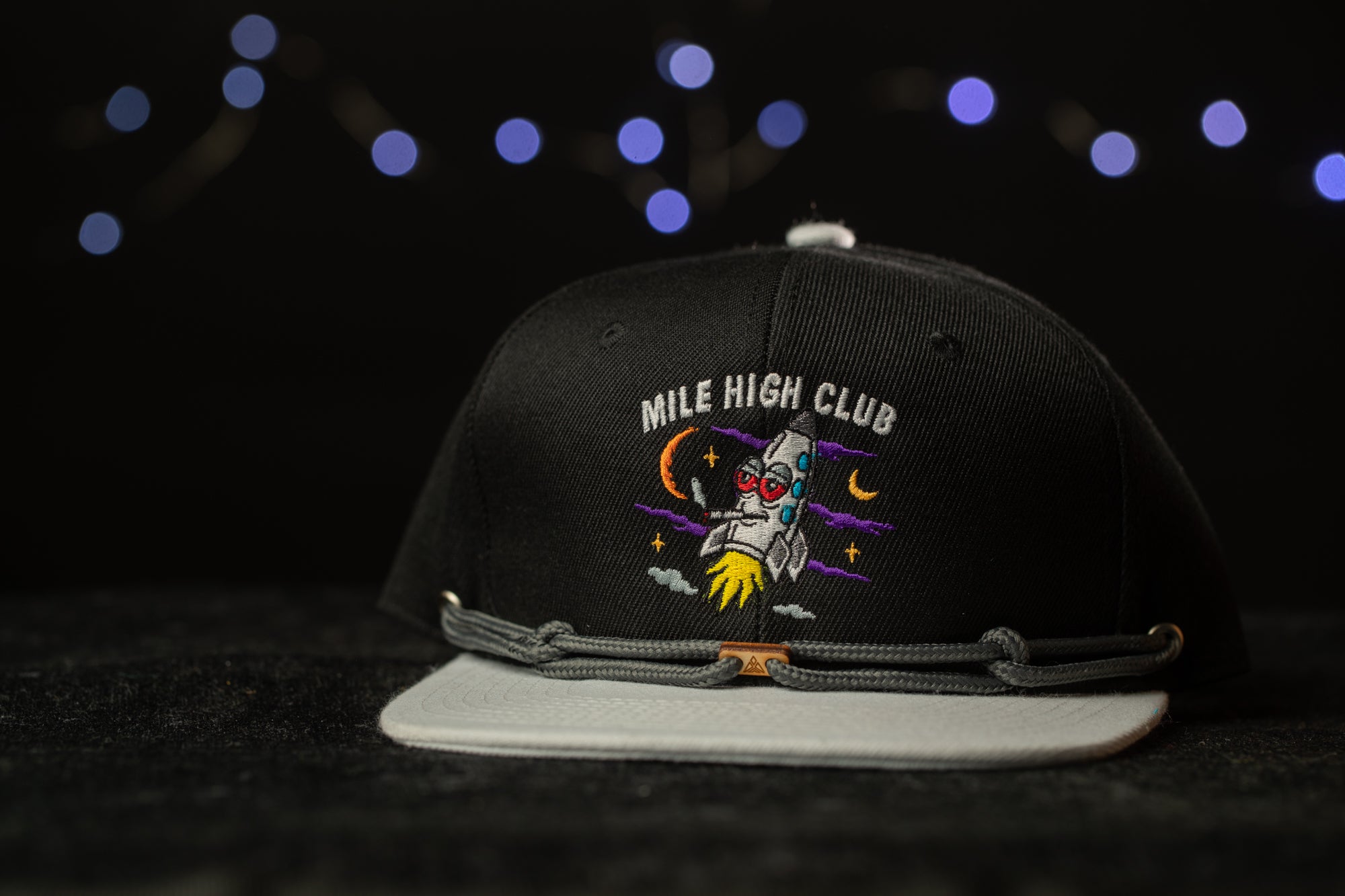 A black baseball cap with a colorful graphic and text on it, placed against a dark background with blurred lights.