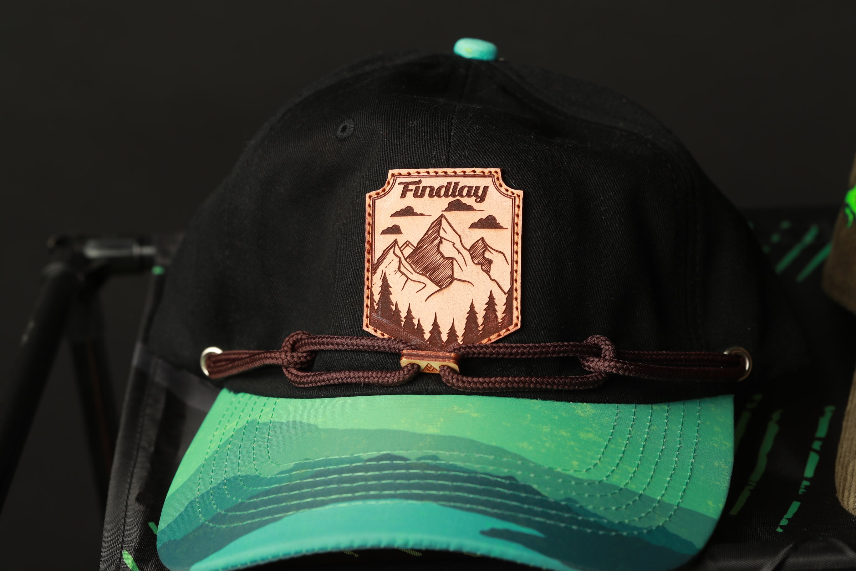 A black baseball cap with a green brim and a brown leather patch featuring a mountain landscape and the word "Findlay" printed on it.