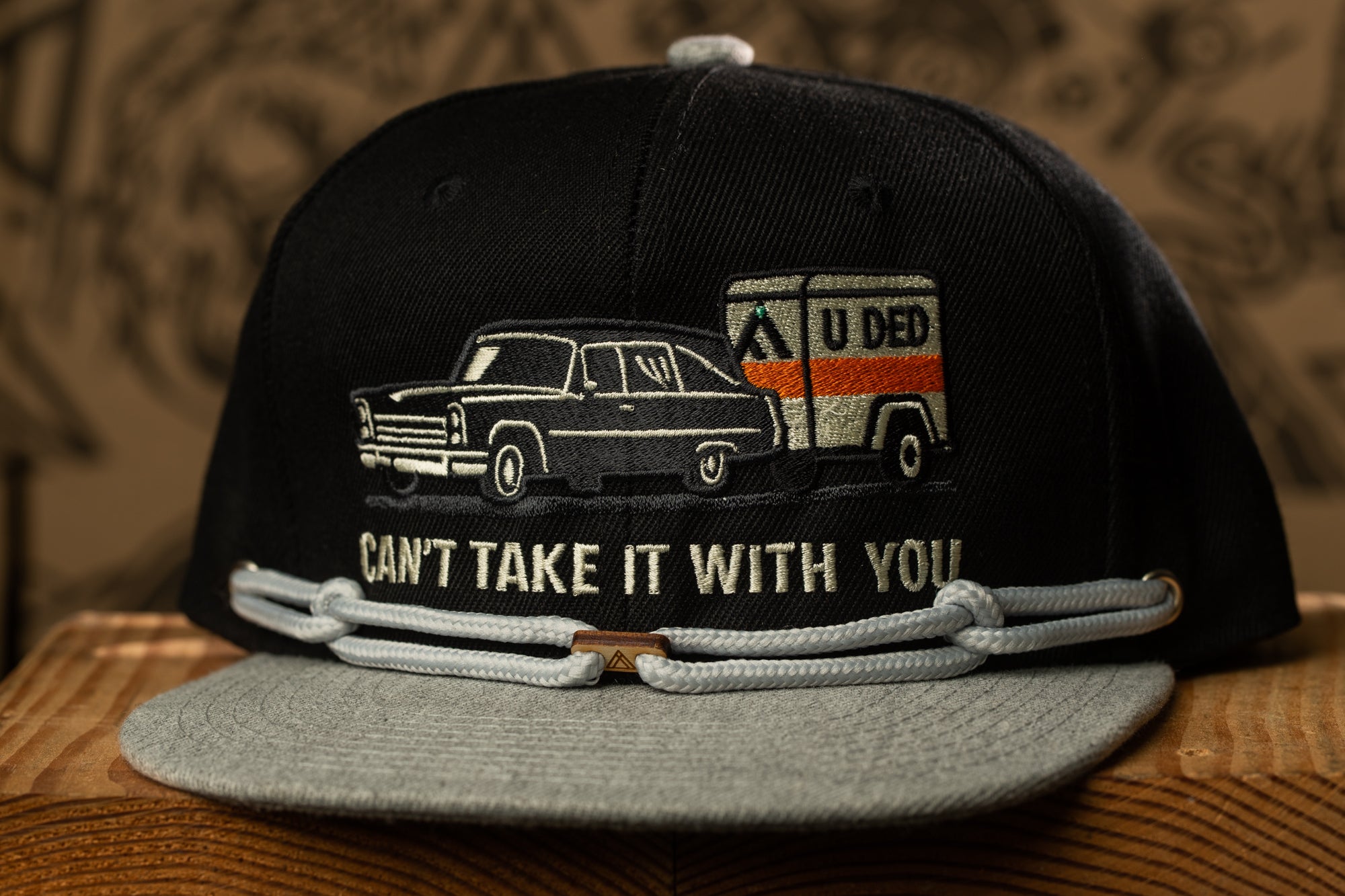 A black baseball cap with a graphic design depicting a car and a trailer, along with the text "CAN'T TAKE IT WITH YOU".