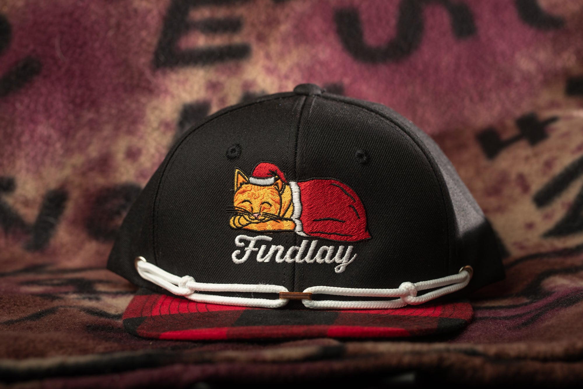 A black baseball cap with a cat embroidered on it, wearing a red Santa hat, and the word "Findlay" written on it.