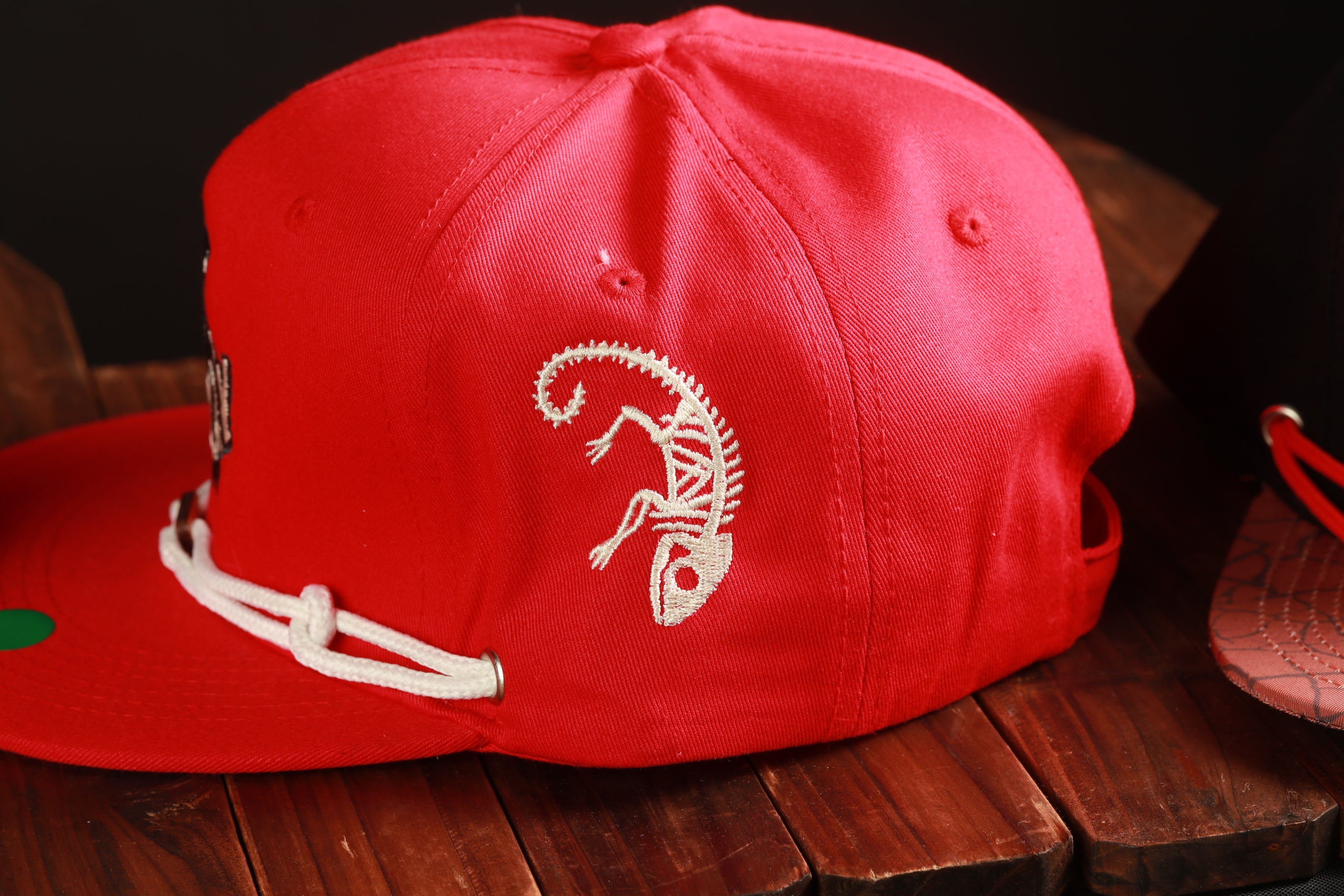 A red baseball cap with a white chameleon design on the side, sitting on a wooden surface.