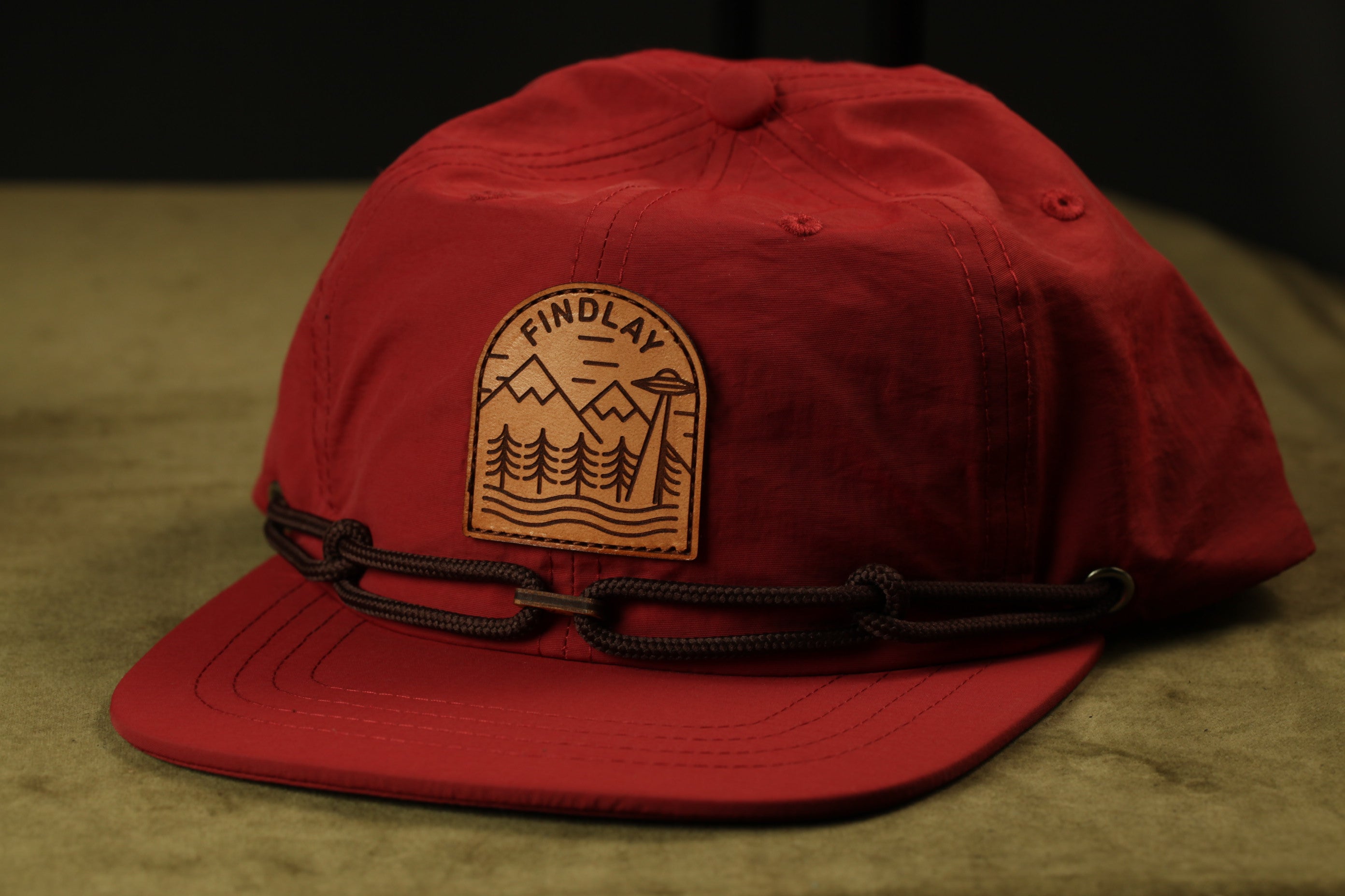 A red baseball cap with a leather patch on the front, featuring a mountain landscape design.
