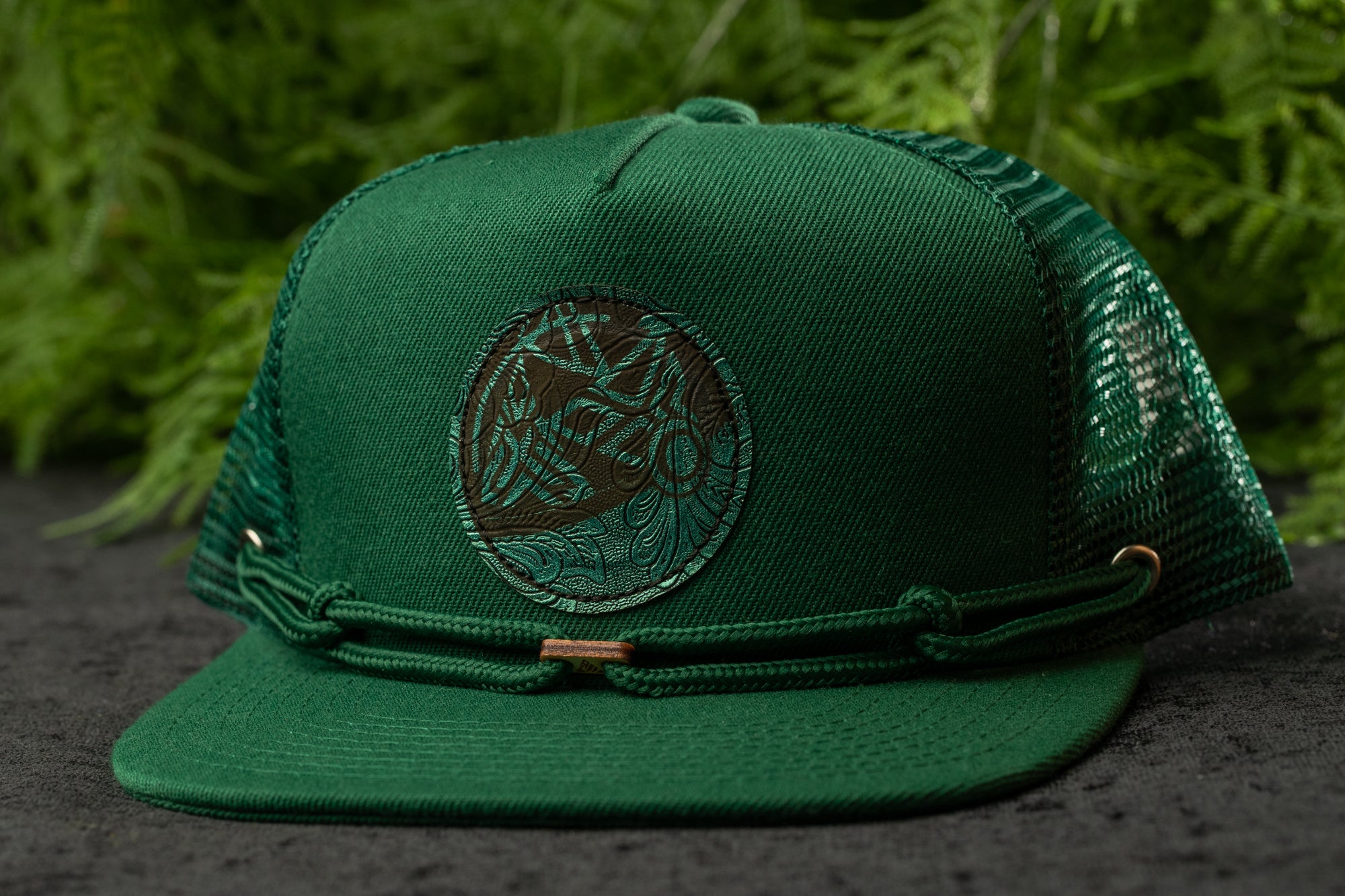 A green baseball cap with a circular logo on the front, placed on a surface with greenery in the background.