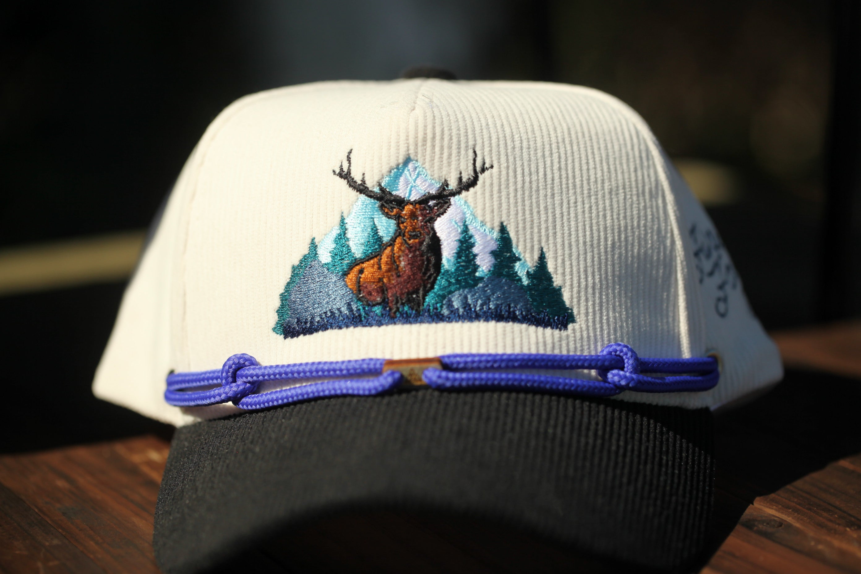 A white baseball cap with a detailed embroidered design of a deer standing in a forest on the front.