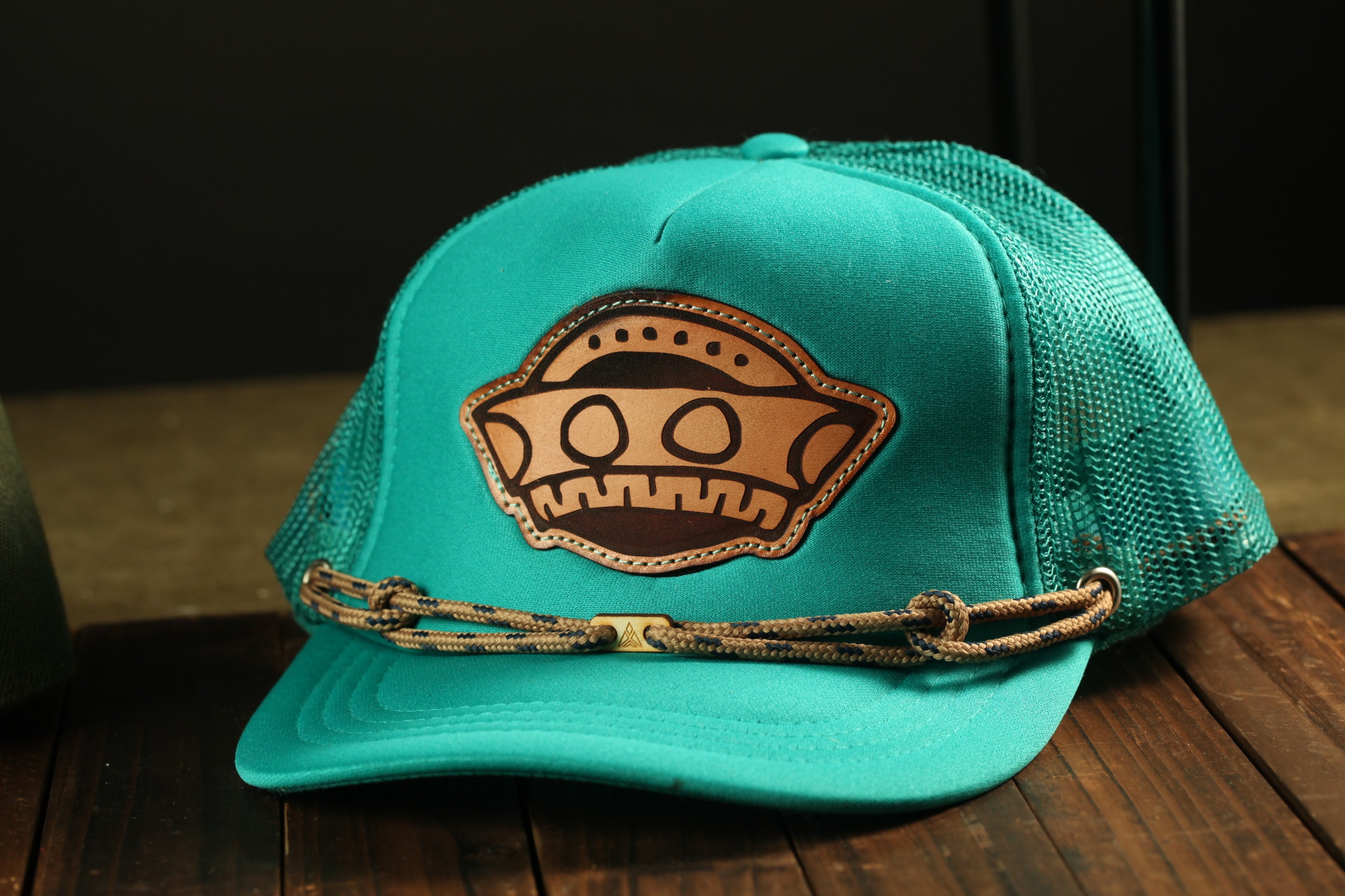 A turquoise baseball cap with a brown leather patch featuring a cartoon-like face design.