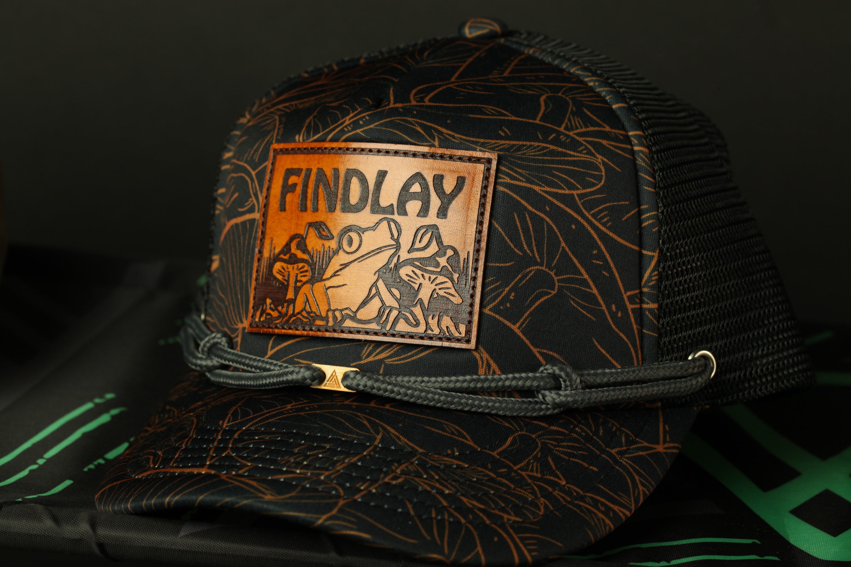A black baseball cap with a leather patch on the front, featuring an illustration of a frog and the text "FINDLAY".