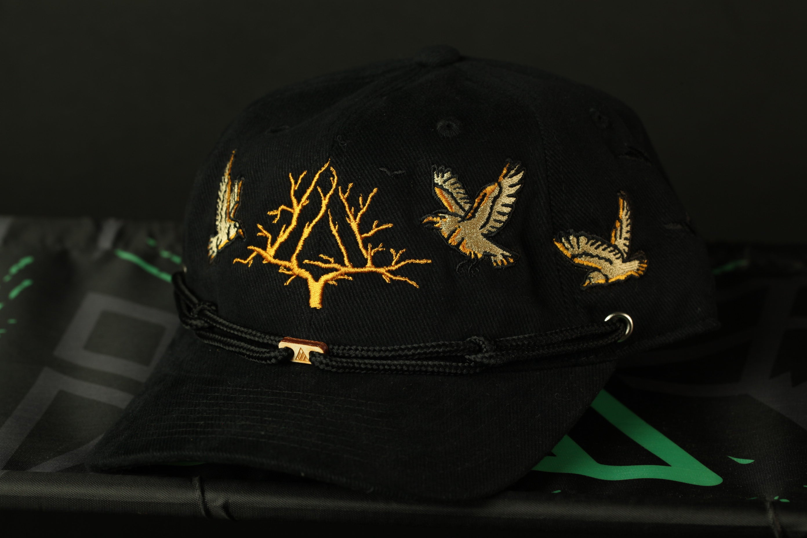 A black baseball cap with gold embroidered designs, including a tree and birds, is shown against a dark background.