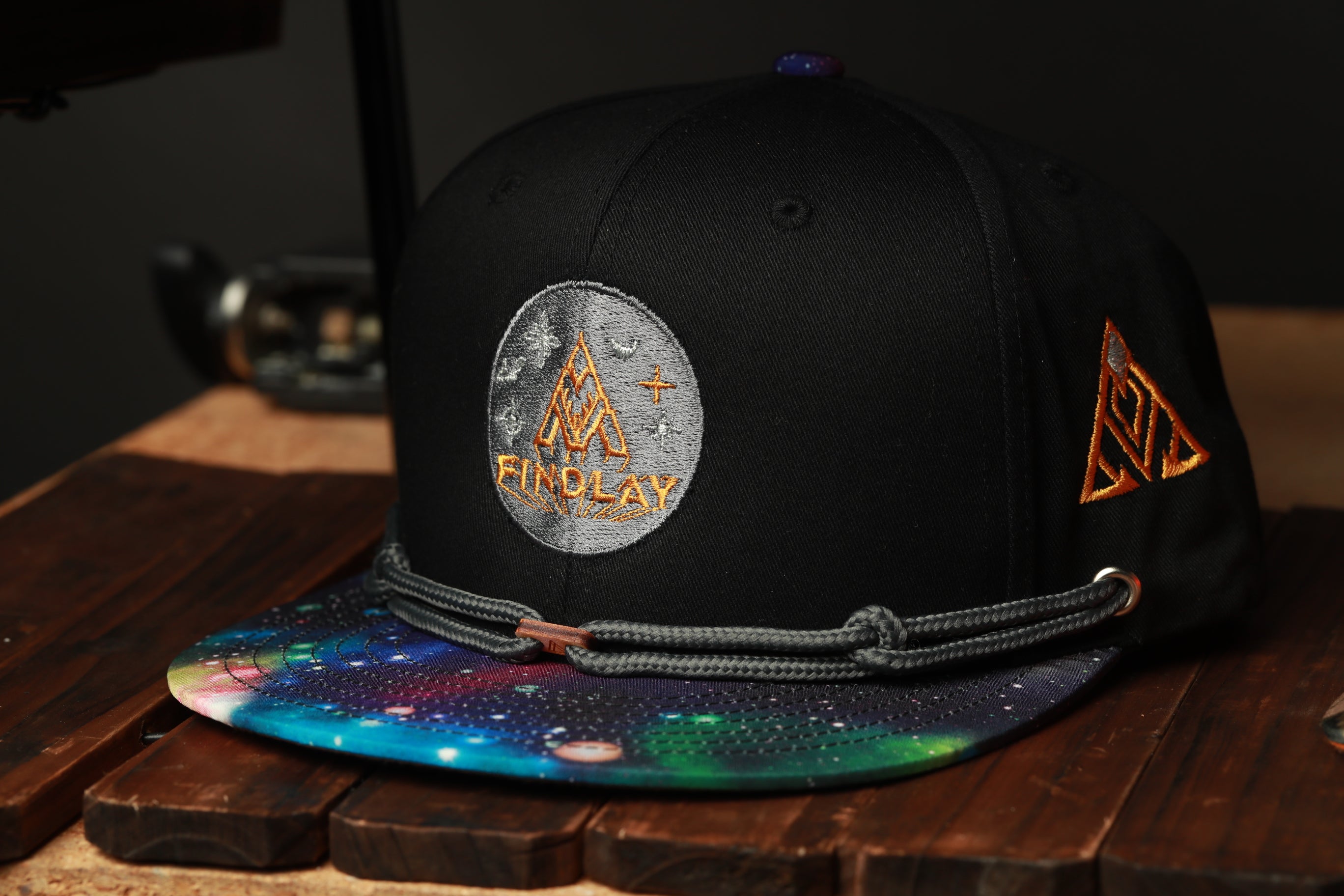 A black baseball cap with a colorful, space-themed design and the word "FINPLAY" printed on it, sitting on a wooden surface.