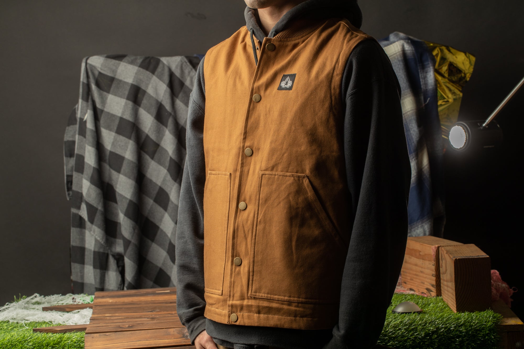 A person wearing a brown vest stands in front of a wooden table with a checkered blanket draped over it.