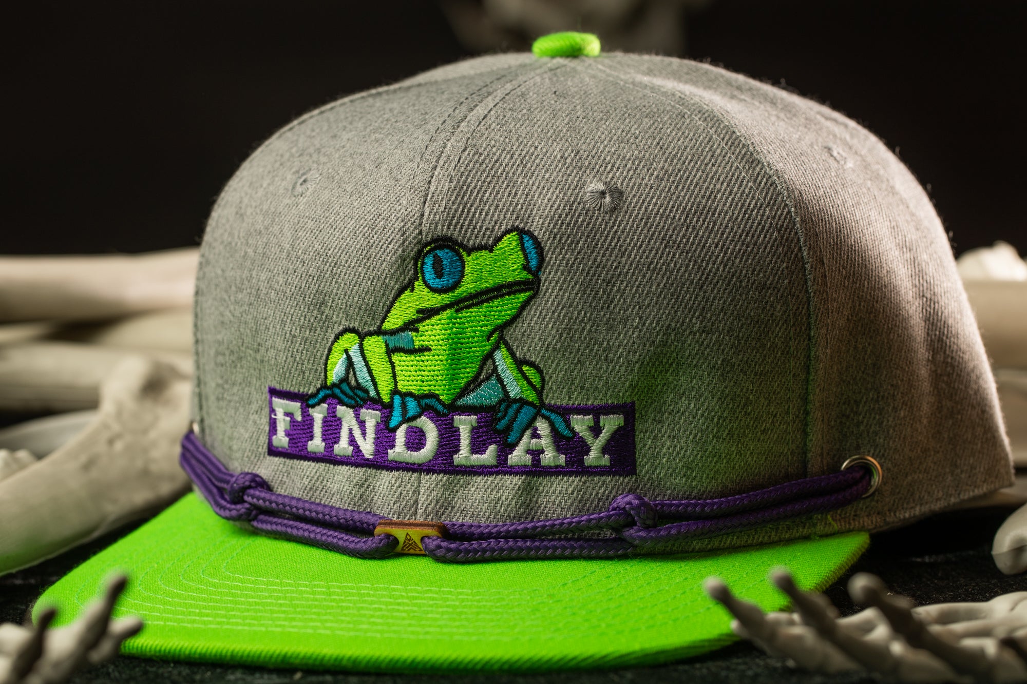 A grey baseball cap with a green frog design and the word "FINDLAY" printed on it, along with a purple band around the brim.
