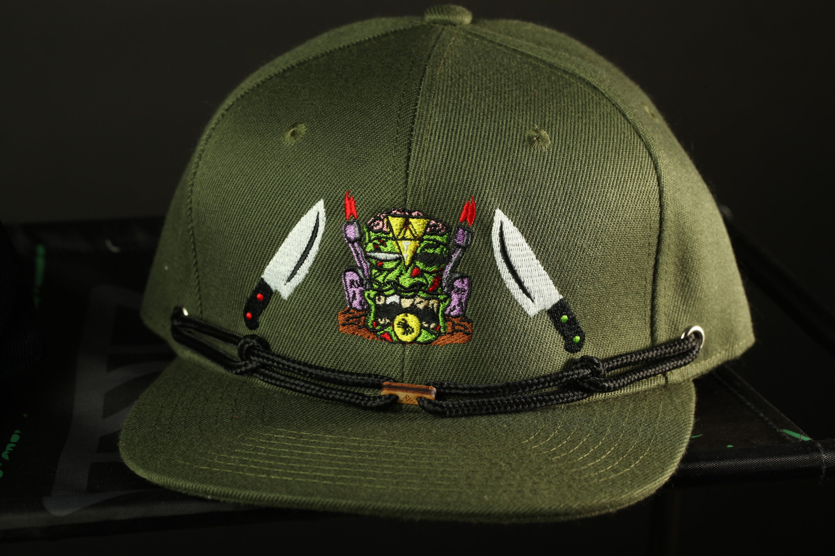 A green baseball cap with a unique design featuring a mask, knives, and a green and yellow color scheme.