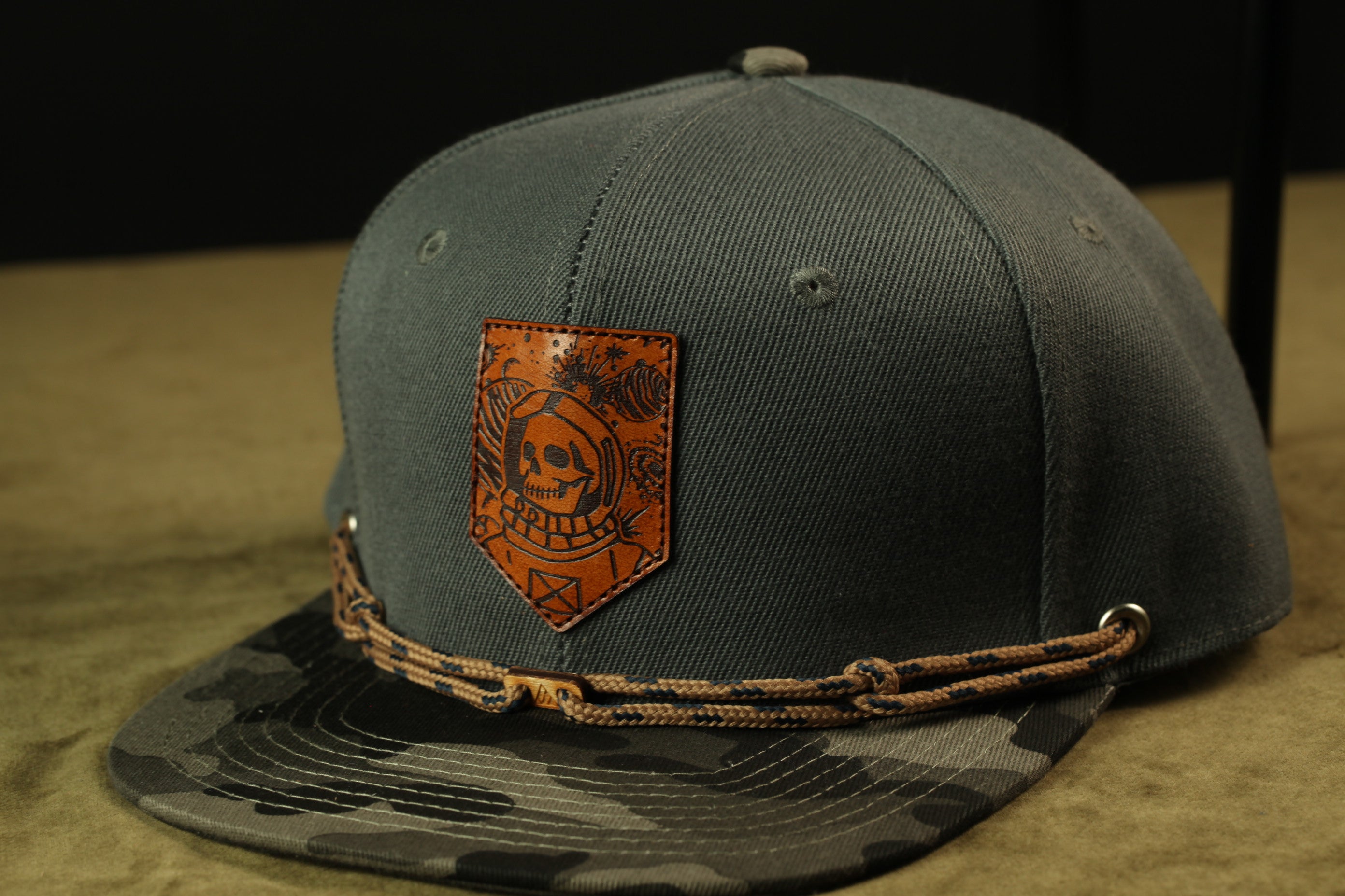 A gray baseball cap with a skull and crossbones patch and a camouflage pattern on the brim.