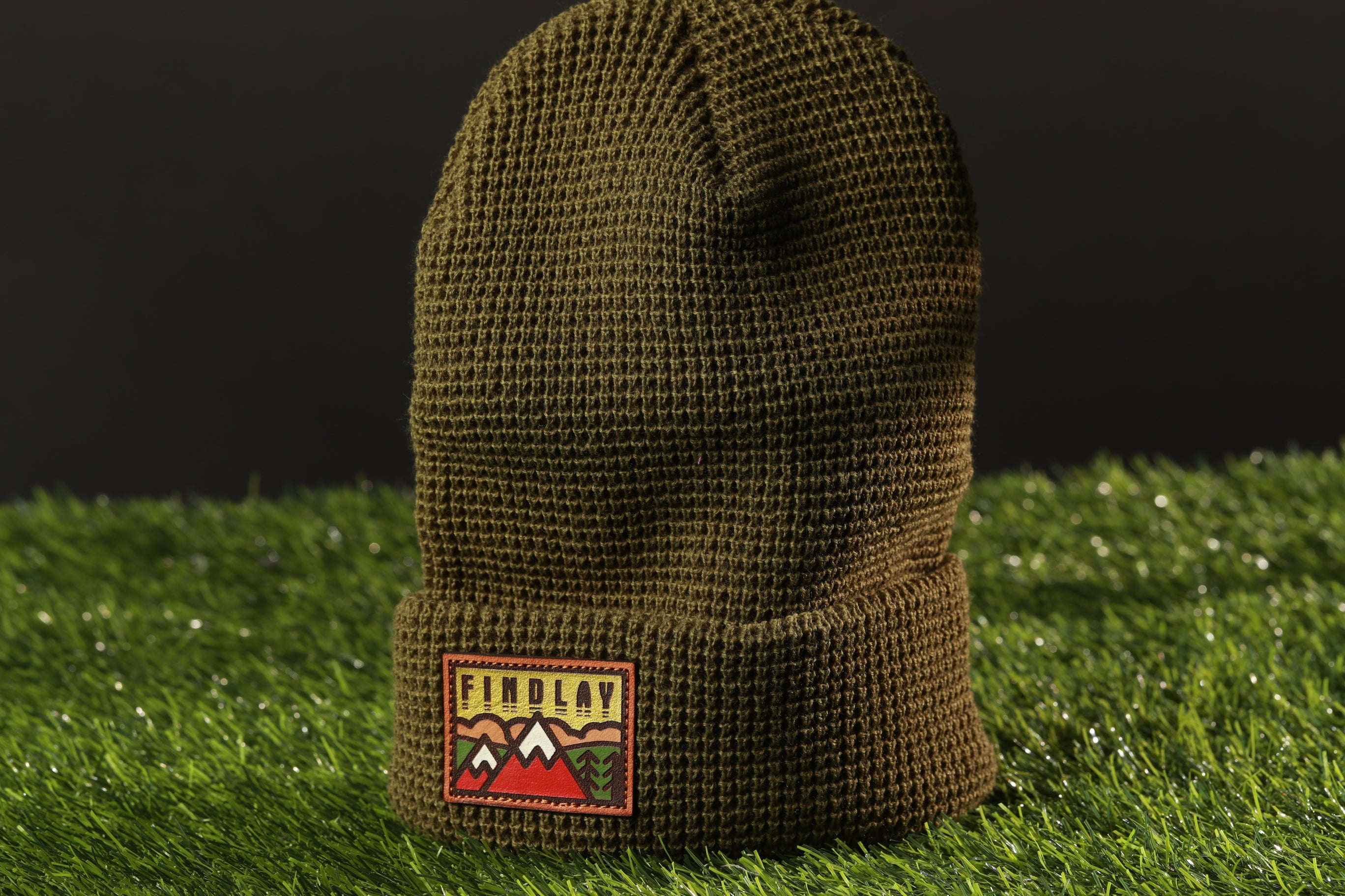 A brown knit beanie with a patch on the front, sitting on a grassy surface.