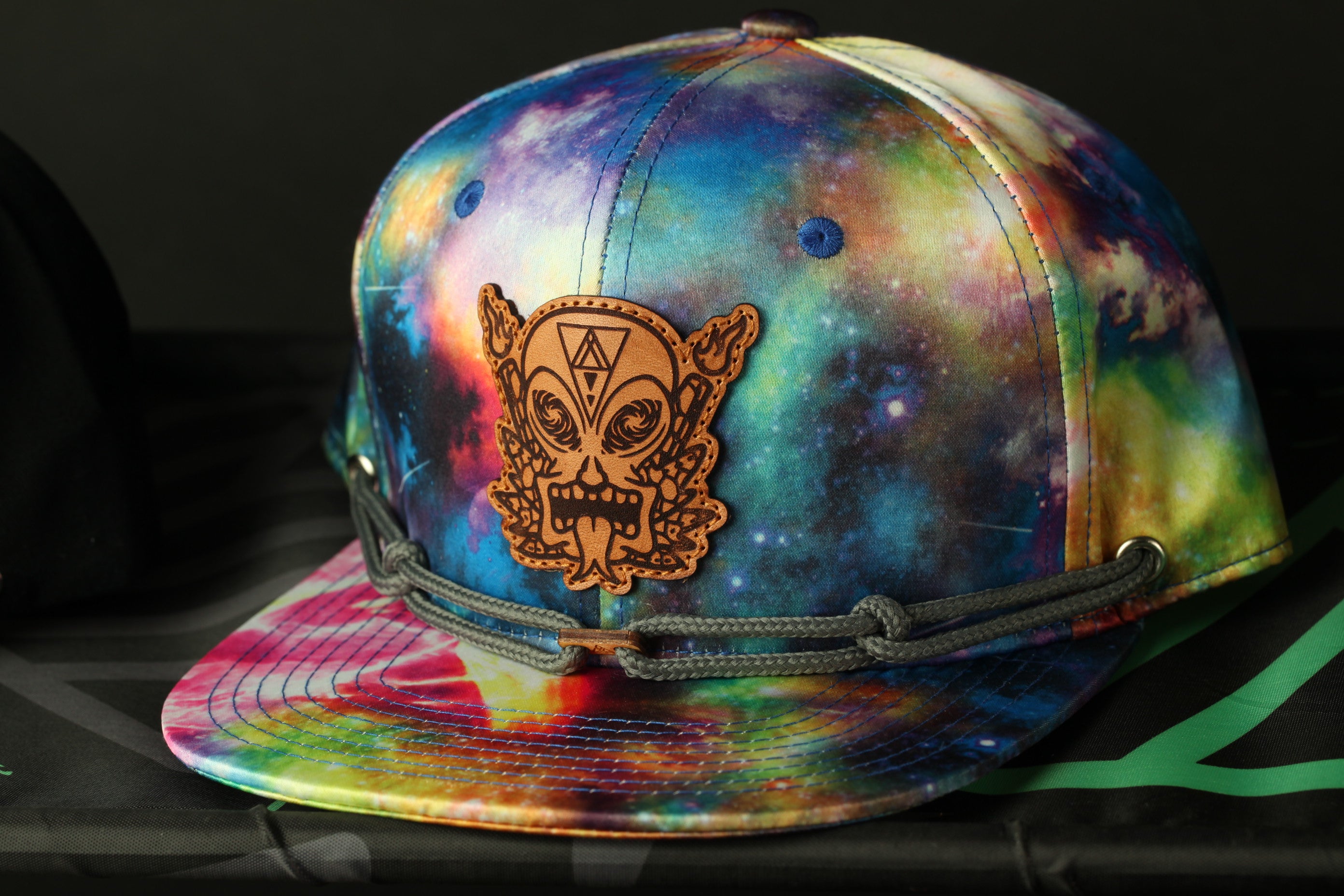 A colorful tie-dye baseball cap with a wooden patch featuring an alien or alien-like face design.