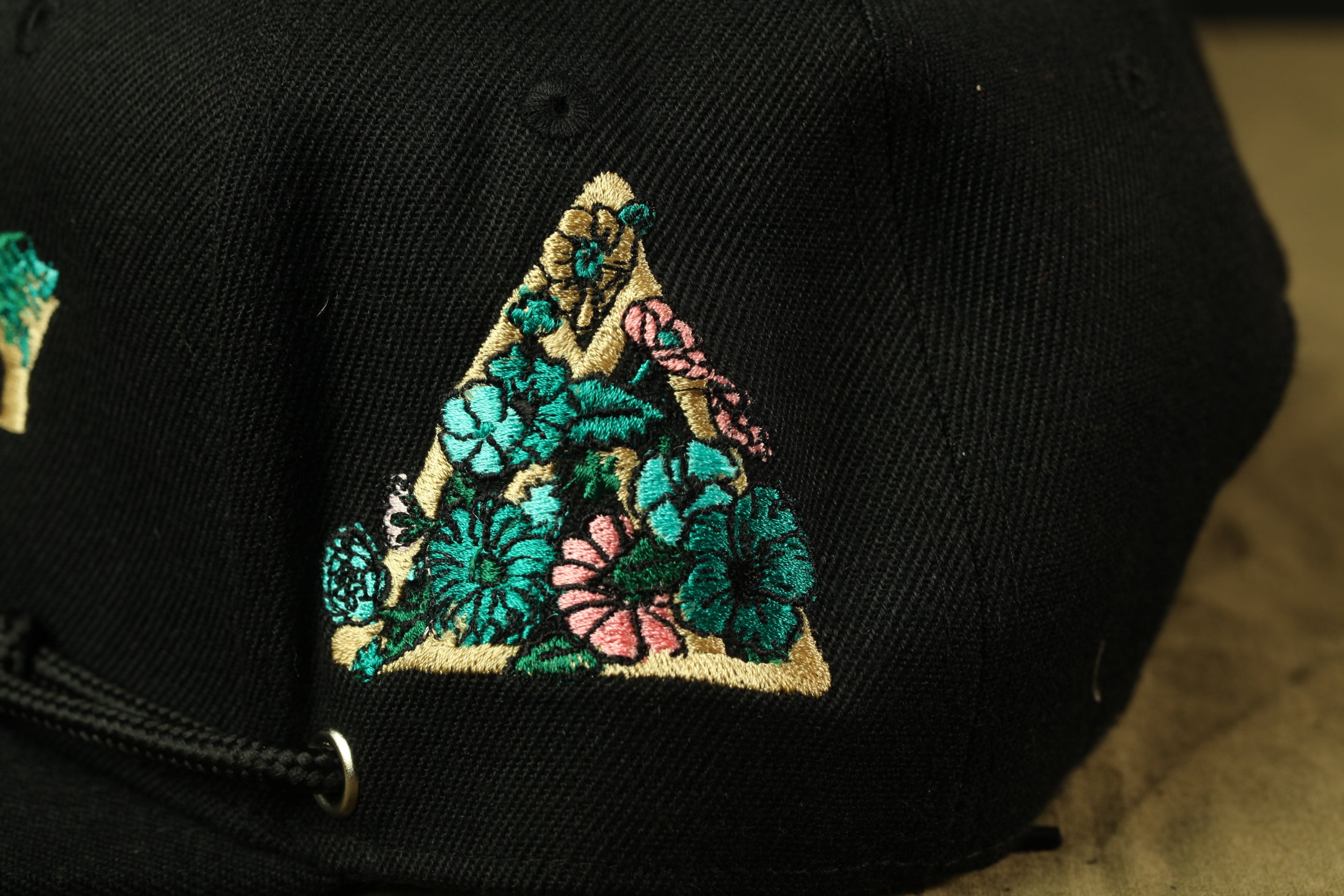 A close-up view of a black baseball cap with a colorful floral embroidered triangle design on the front.