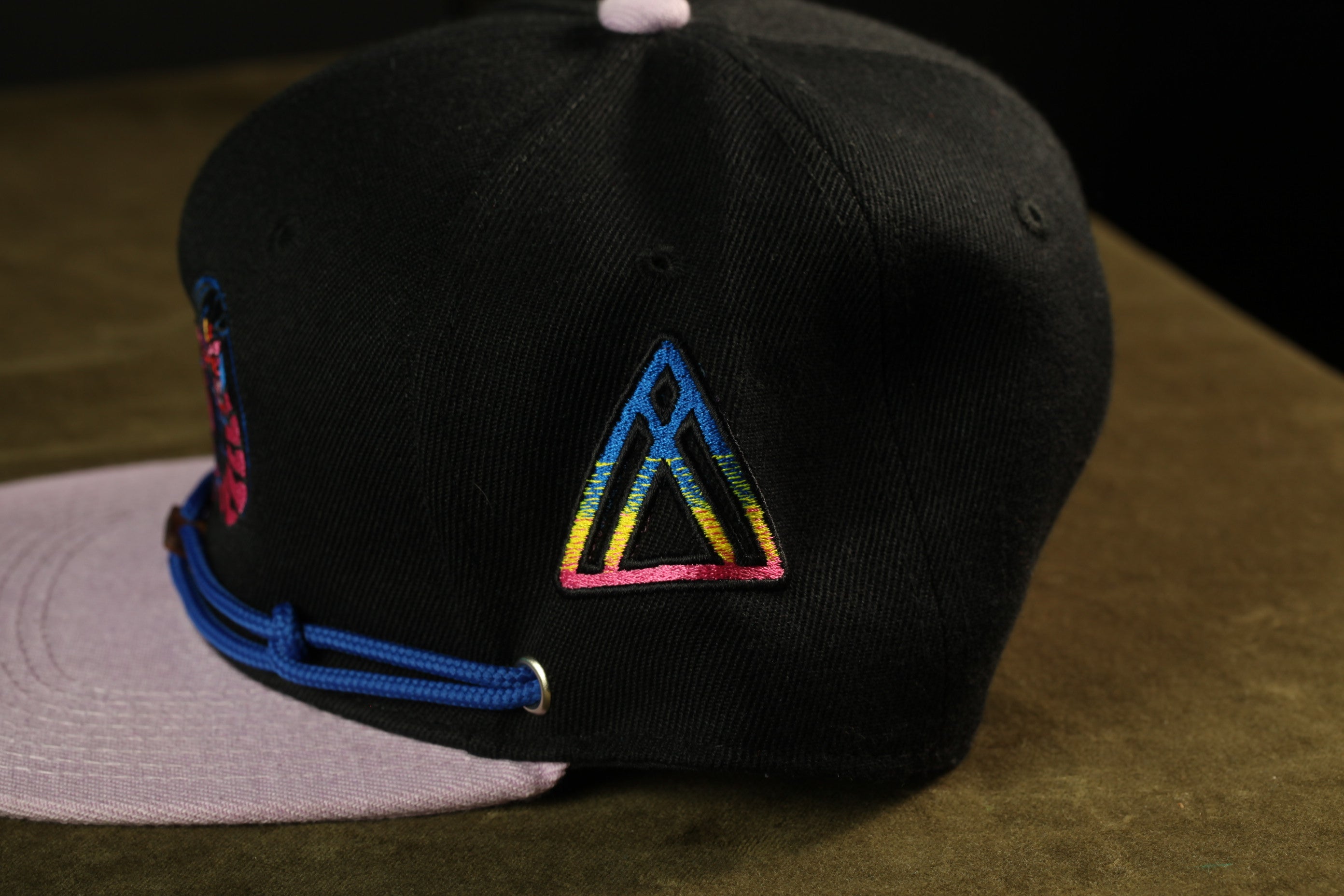 A black baseball cap with a colorful triangle-shaped logo on the front, resting on a brown surface.